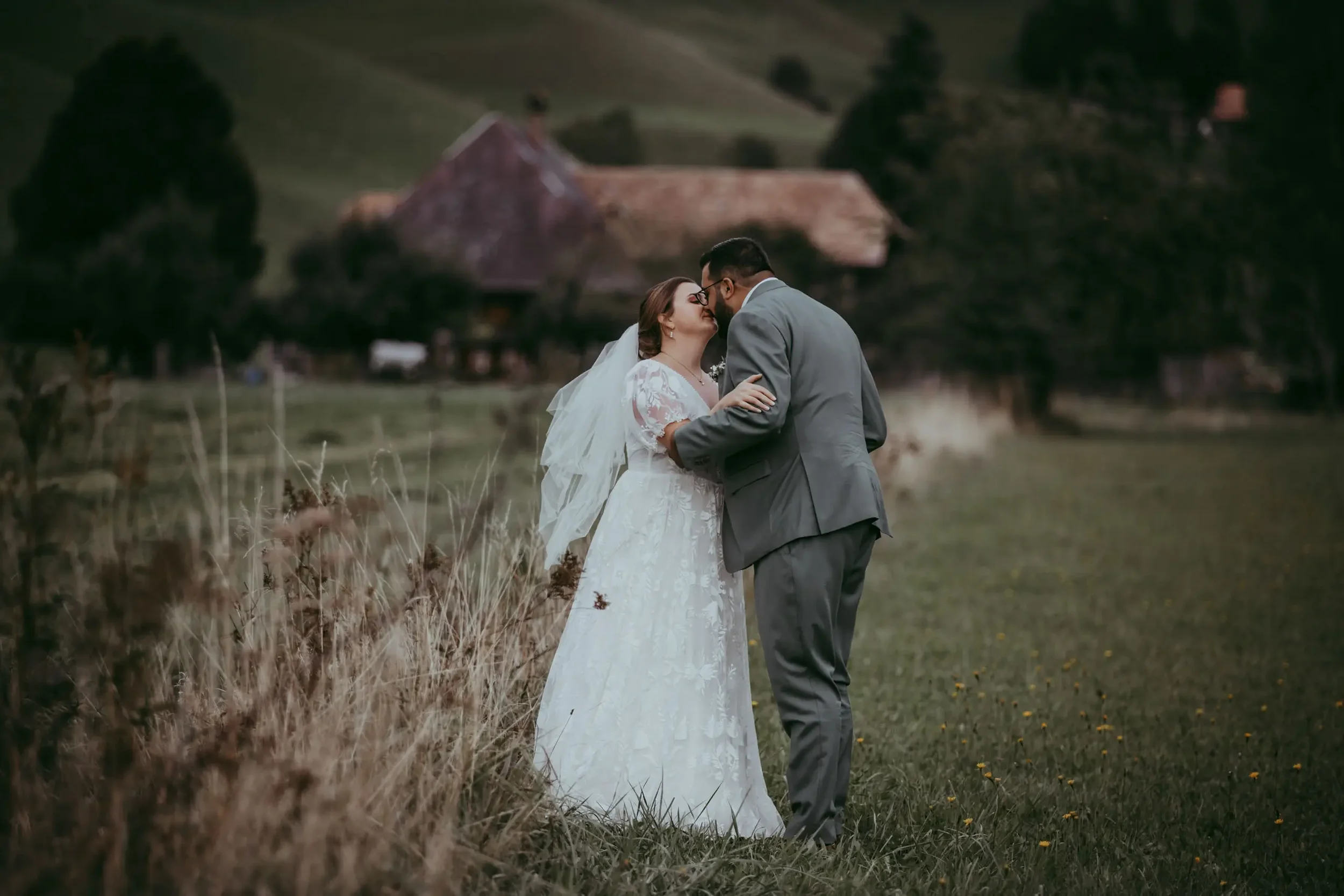Ein Brautpaar steht im Grünen, umgeben von Feldern und Bäumen, bei einer ländlichen Hochzeit, küsst sich. Im Hintergrund ist ein Haus zu sehen.