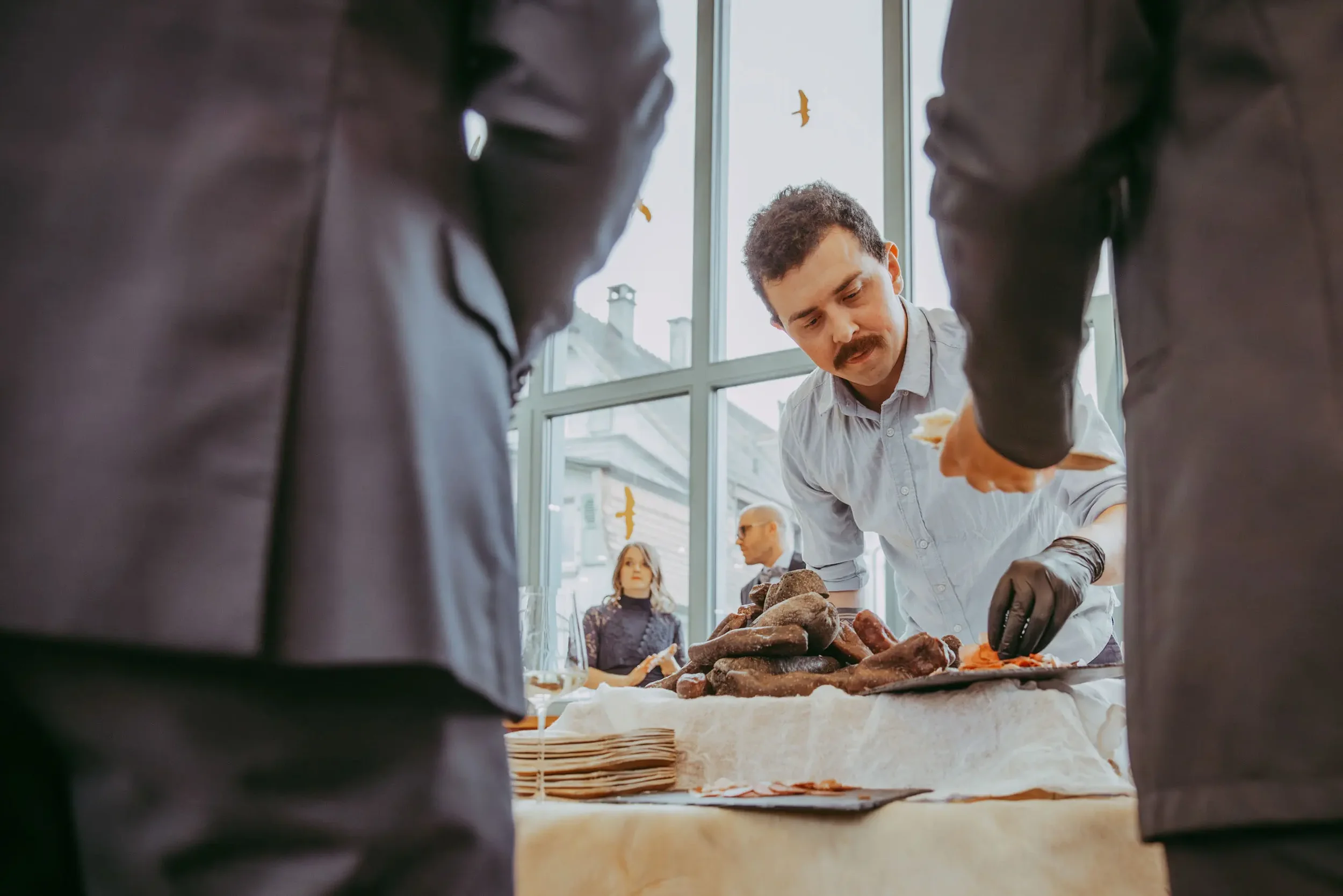 Ein Mann beim Verteilen von Brot auf einem Buffet, im Hintergrund Menschen im Raum, große Fenster mit Blick nach draußen.