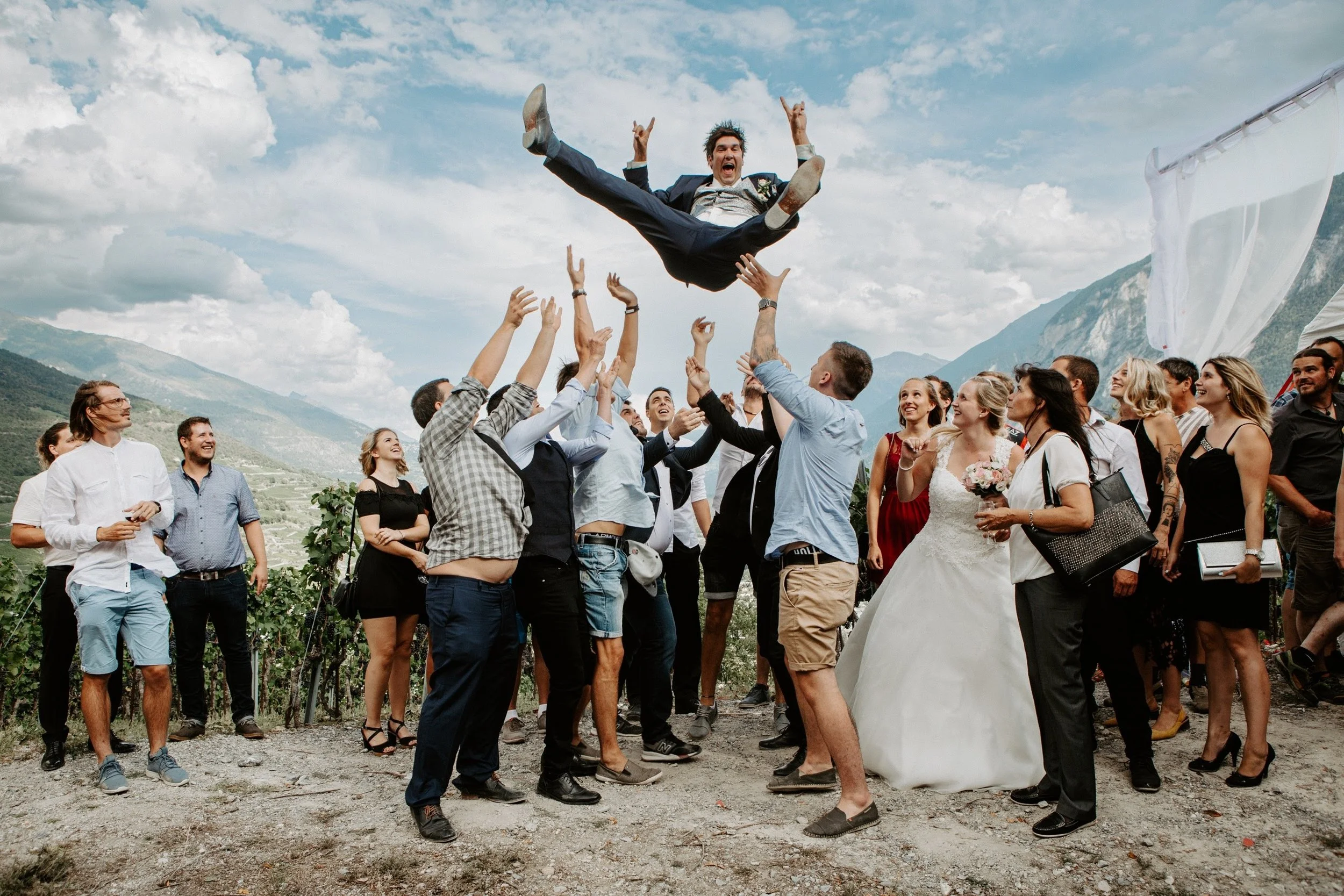 Ein Mann in einem Anzug wird bei einer Feier von einer Gruppe von Menschen hochgehoben, im Hintergrund Berge und ein blauer Himmel mit Wolken.