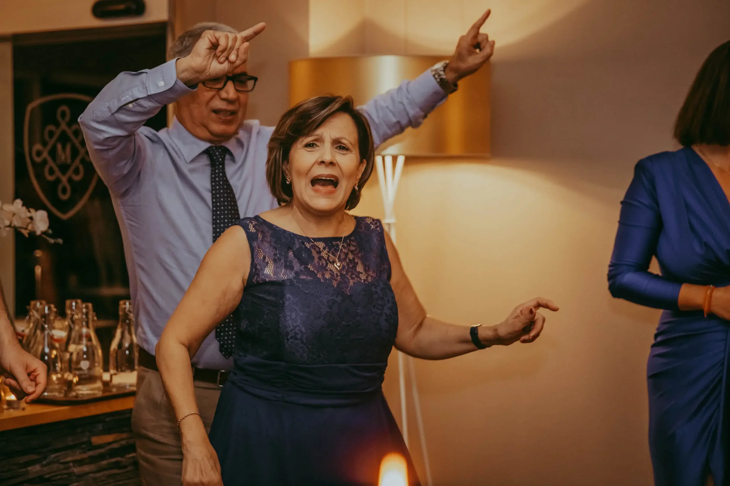 Zwei Menschen, ein Mann und eine Frau, scheinen in einer Party oder einem Fest zu tanzen oder zu feiern, im Hintergrund ein Tisch mit Getränken und eine Stehlampe.