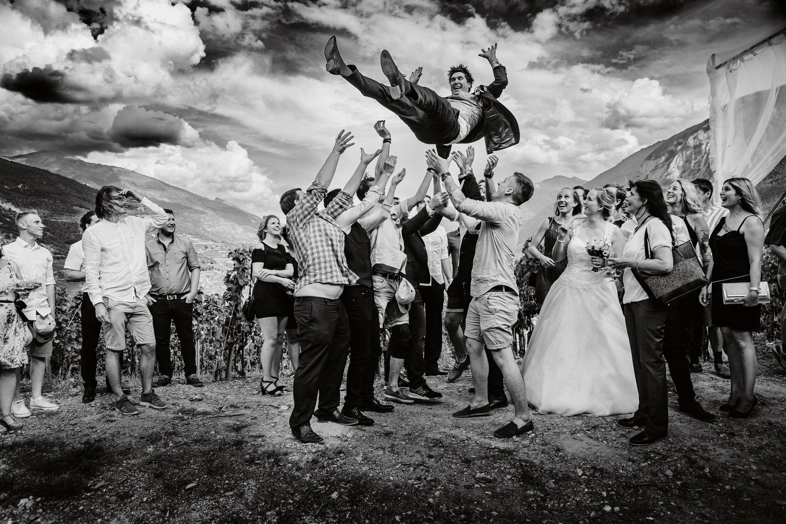 Schwarz-Weiß-Foto einer Hochzeit im Freien, bei der ein Mann in Anzug und Jacke in die Luft gehoben wird, umgeben von vielen lachenden Menschen, die an einer Hochzeit teilnehmen. Im Hintergrund sind Berge und Wolken sichtbar.