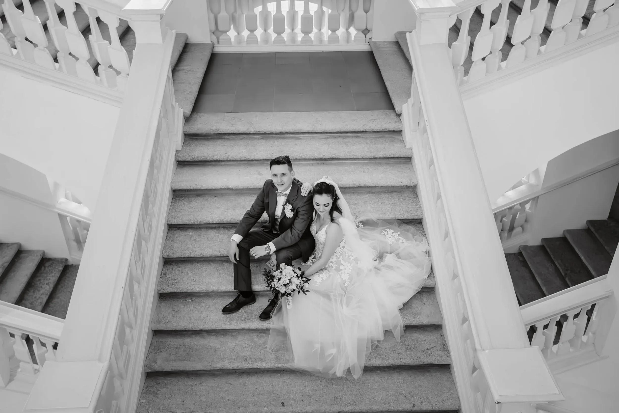 Braut und Bräutigam sitzen auf einer Treppe in einem eleganten Gebäude, vermutlich nach ihrer Hochzeit, umrahmt von weißen Geländern.