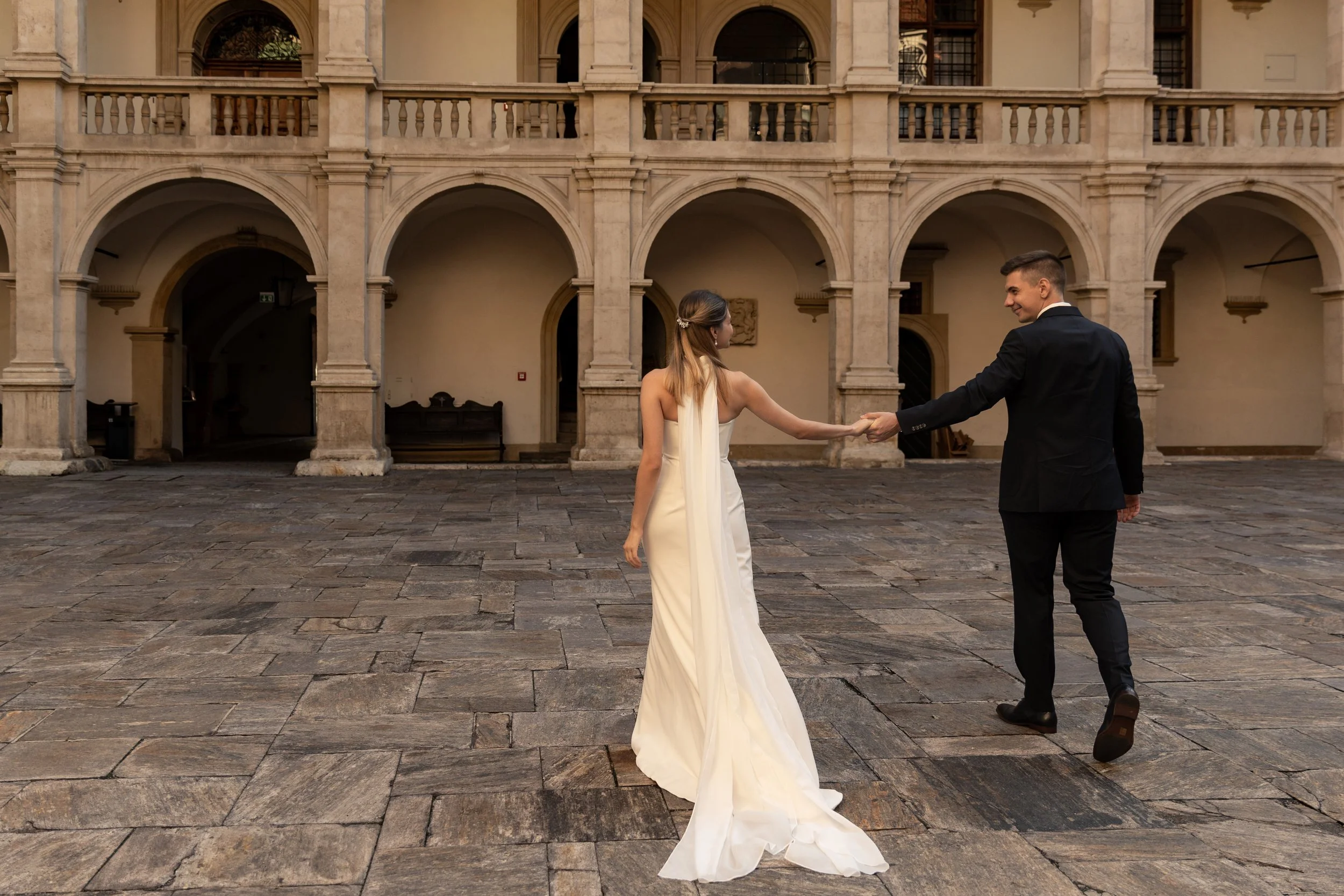 Ein Mann in Anzug hält die Hand einer Frau in einem weißen Brautkleid in einem historischen Innenhof mit Arkaden und Steinböden.