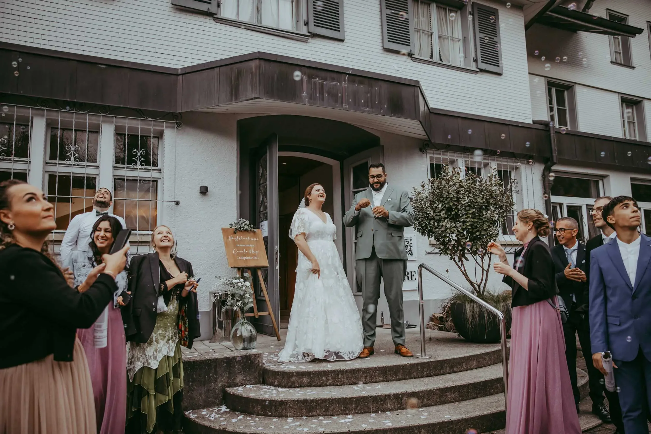 Hochzeitspaar auf Treppe vor einem Haus mit mehreren Gästen, die zuschauen und Fotos machen, bei Sonnenschein.