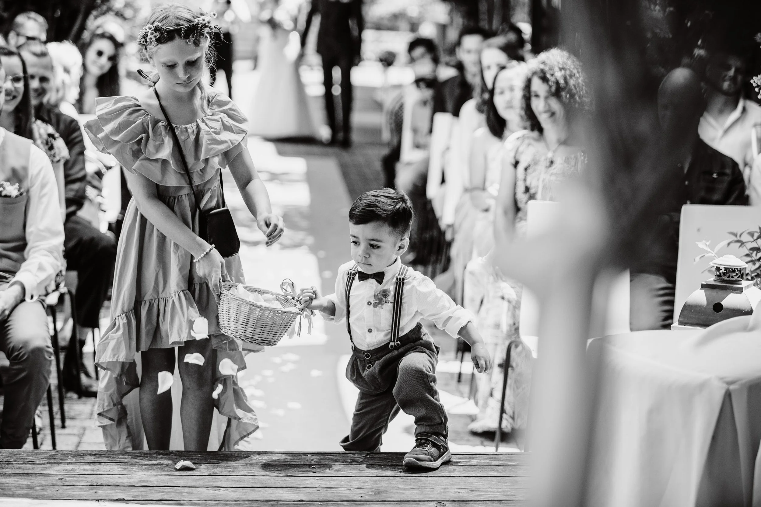 Schwarz-Weiß-Foto eines Kindergeburtstags im Freien mit sitzenden Erwachsenen und einem Jungen, der mit Ballons spielt, während eine Frau im Kostüm den Ballons nachjagt.