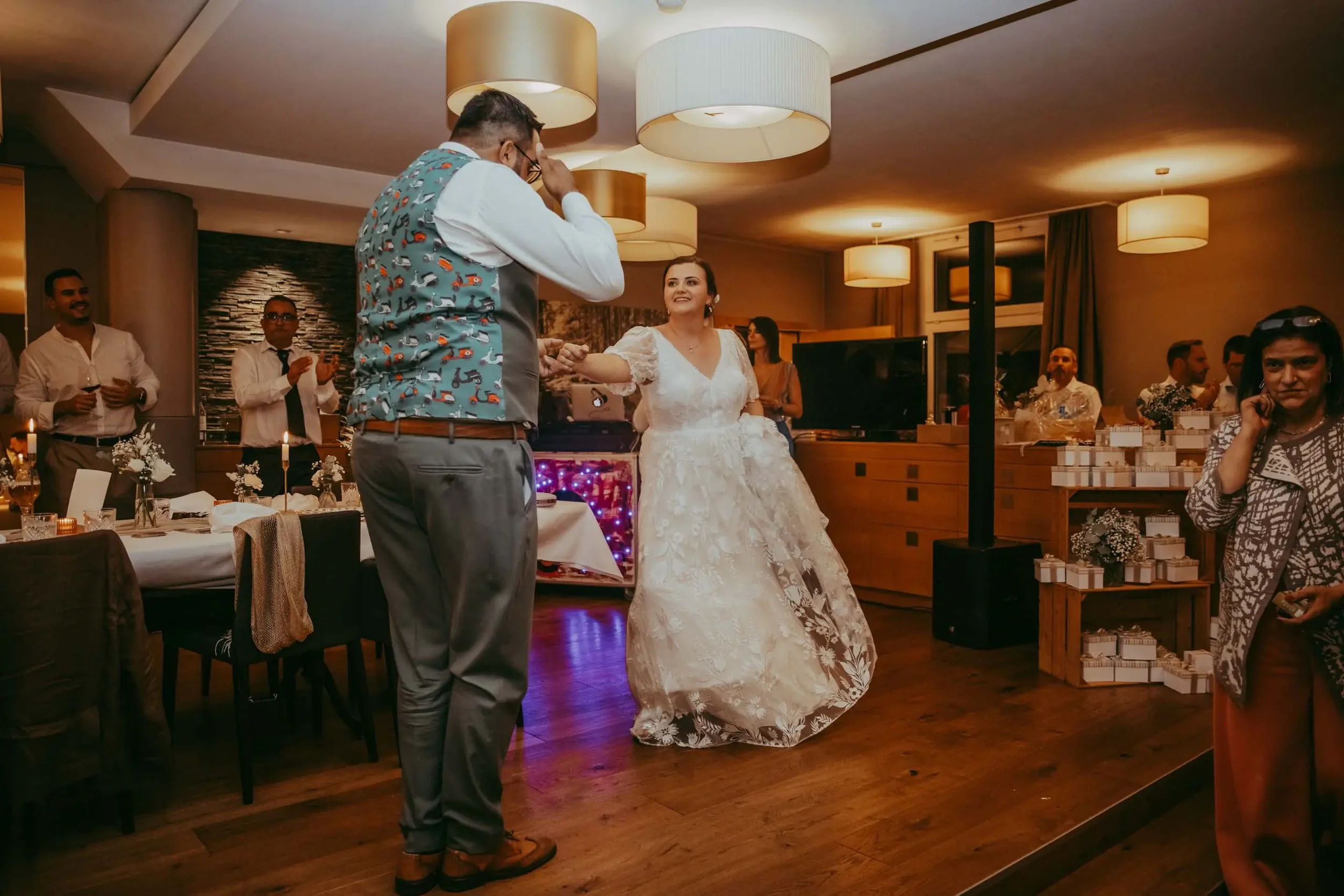 Ein Brautpaar tanzt auf einer Hochzeit. Die Braut trägt ein weißes Brautkleid, der Bräutigam trägt ein Hemd und eine Weste mit Motiven. Gäste im Hintergrund feiern in einem festlich dekorierten Raum.