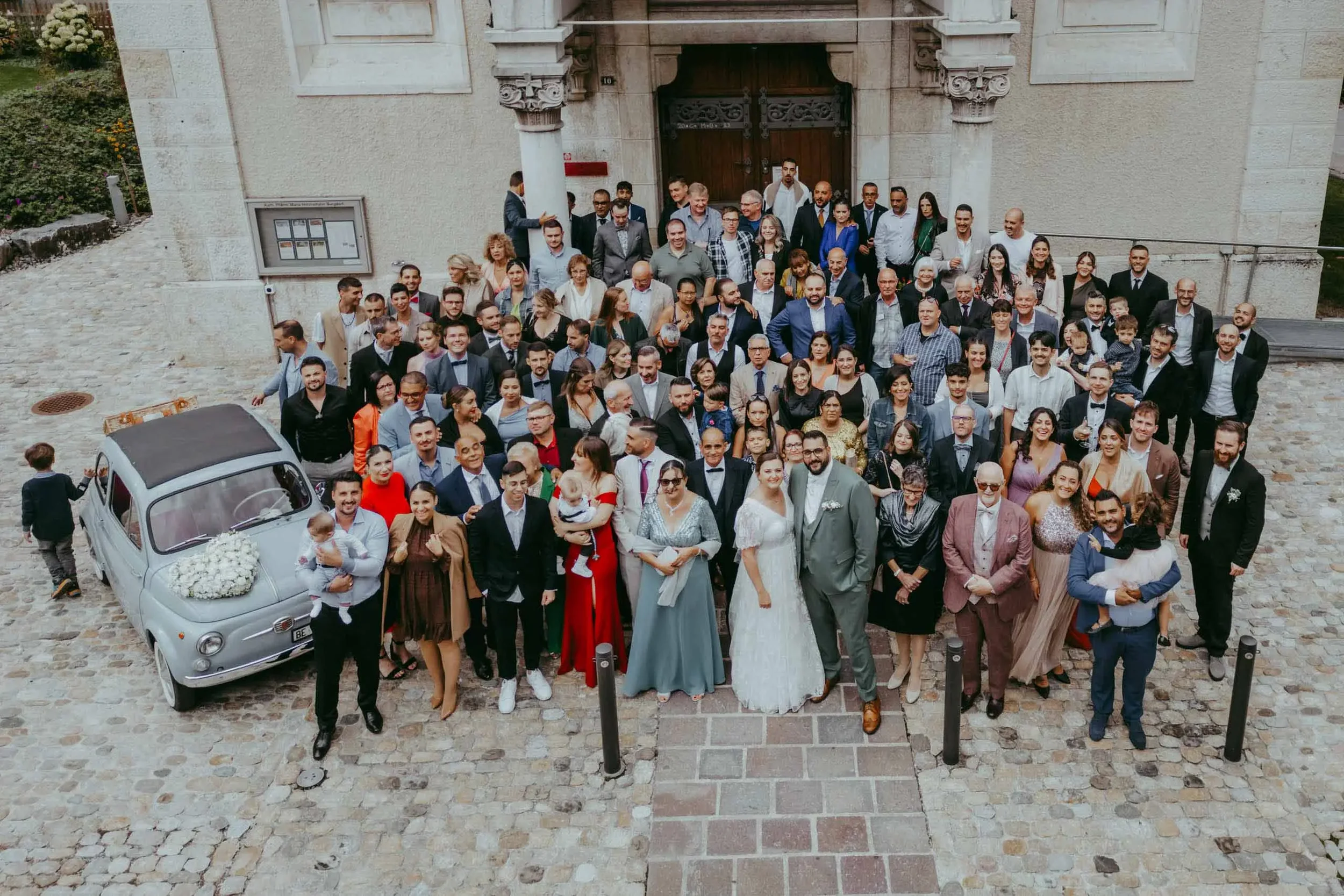 Gruppe von Menschen vor einer alten Gebäudetür bei einer Feier, mit einem Oldtimer im Vordergrund, einige Menschen sind gekleidet wie bei einer Hochzeit.