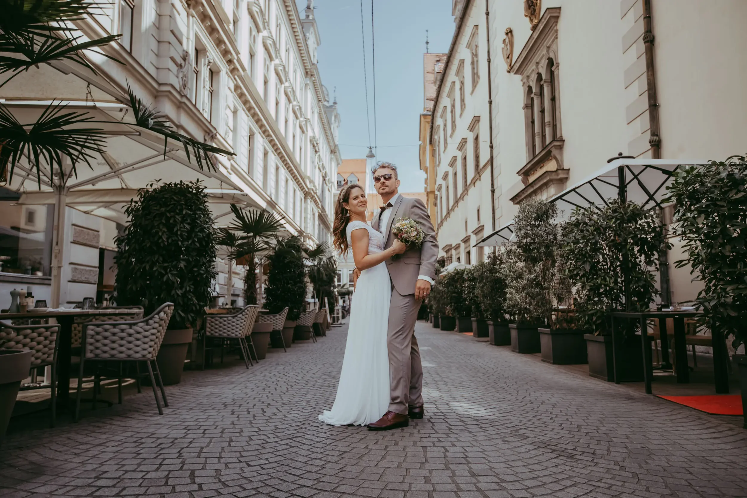 Ein frisch verheiratetes Paar steht auf einer Kopfsteinpflasterstraße in einer urbanen Umgebung, umgeben von Tischen im Freien, Pflanzen und großen Gebäuden mit klassischen Fassaden. Die Frau trägt ein weißes Hochzeitskleid mit einem Bouquet, der Man