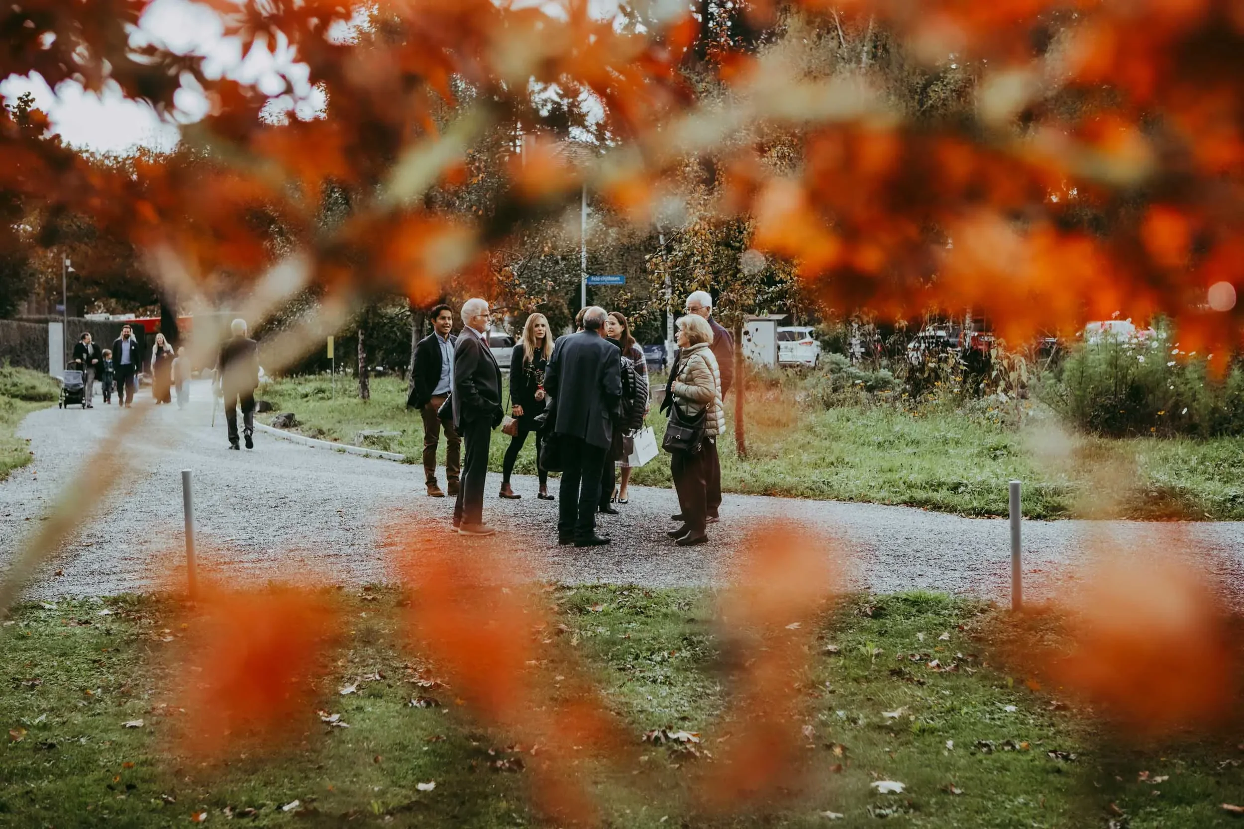 Eine Gruppe von Menschen steht im Gespräch in einem Park, eingefasst durch orangefarbene Herbstblätter im Vordergrund.