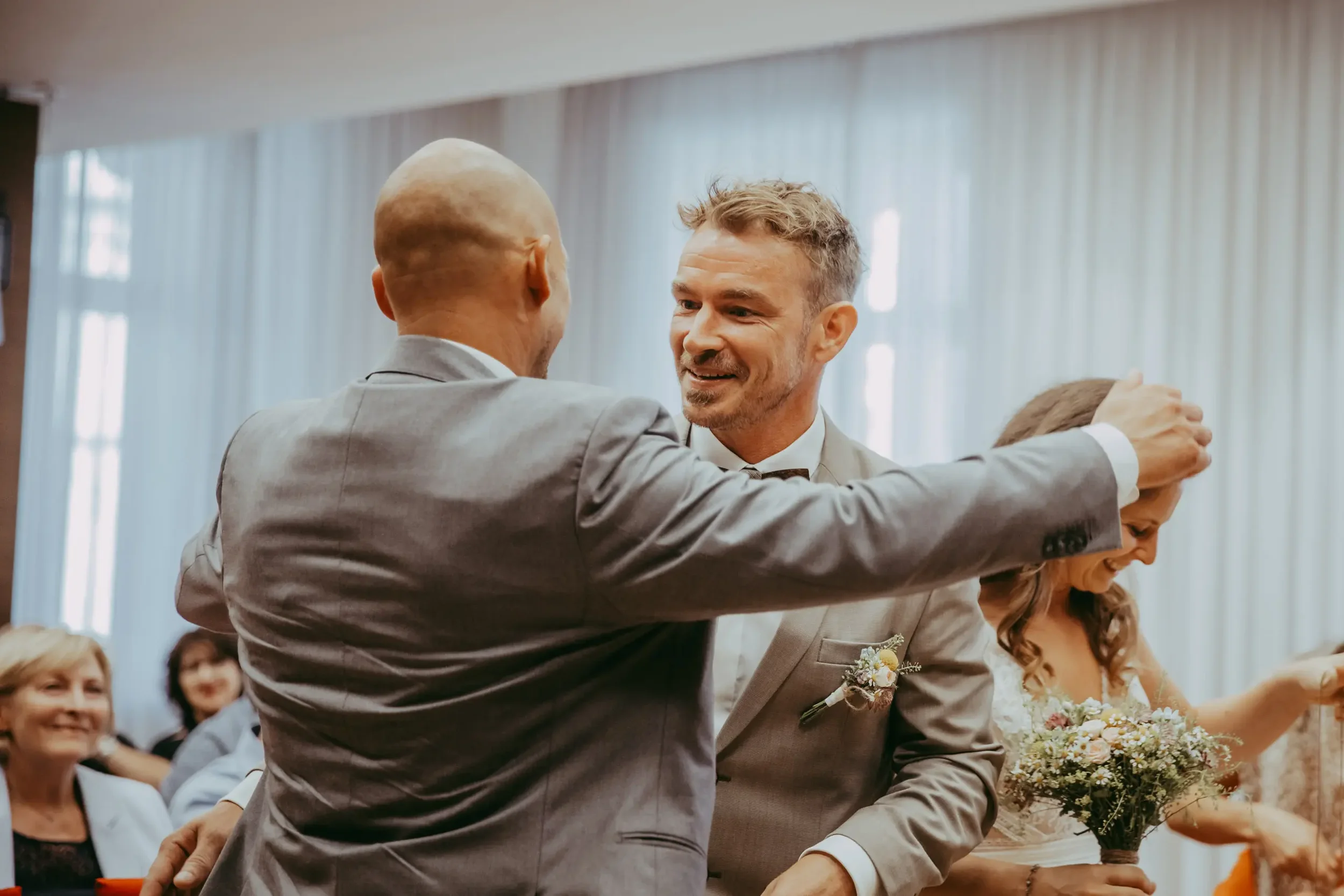 Zwei Männer, die sich auf einer Hochzeit umarmen, während andere Gäste im Hintergrund zuschauen.