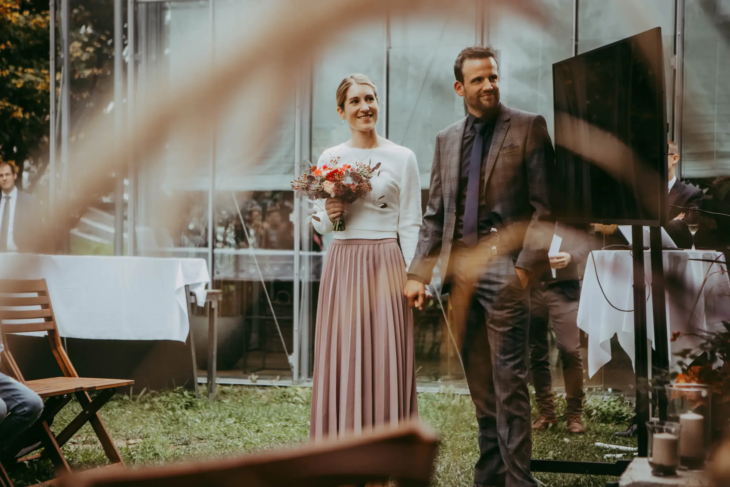 Ein Brautpaar bei einer Hochzeitsfeier im Freien, die Frau hält einen Blumenstrauß, beide lächeln, im Hintergrund sind Tische und Stühle.