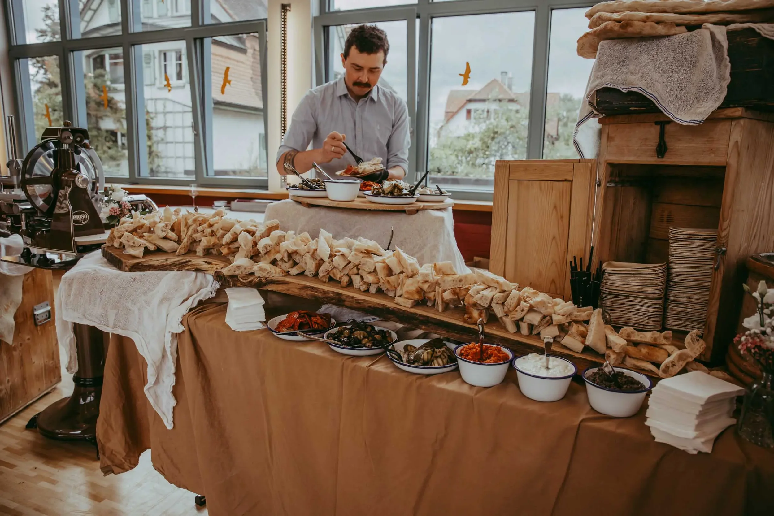 Ein Mann steht hinter einem Buffet mit verschiedenen Speisen, darunter Brot, Aufstriche und verschiedene mediterrane Gerichte, in einem hell erleuchteten Raum mit großen Fenstern.