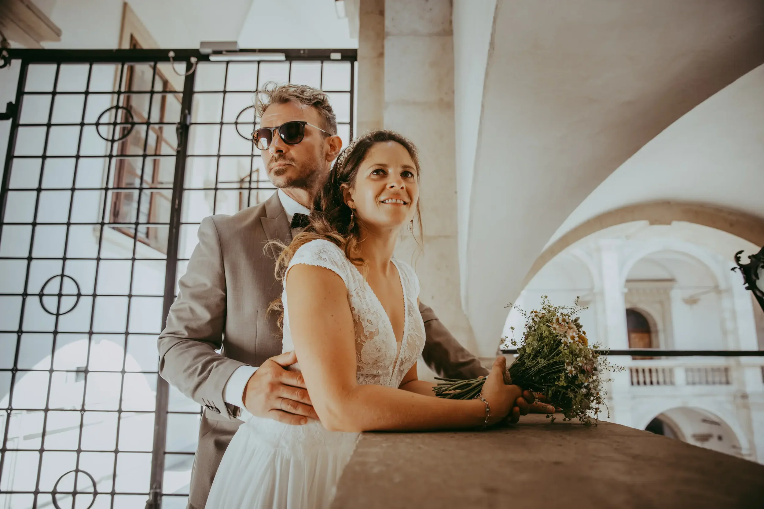 Ein verheiratetes Paar bei einer standesamtlichen Hochzeit in einer historischen, hell erleuchteten Location, mit dem Bräutigam in Anzug und Sonnenbrille, der Braut in einem weißen Kleid mit Blumenstrauß, umarmt am Tisch.