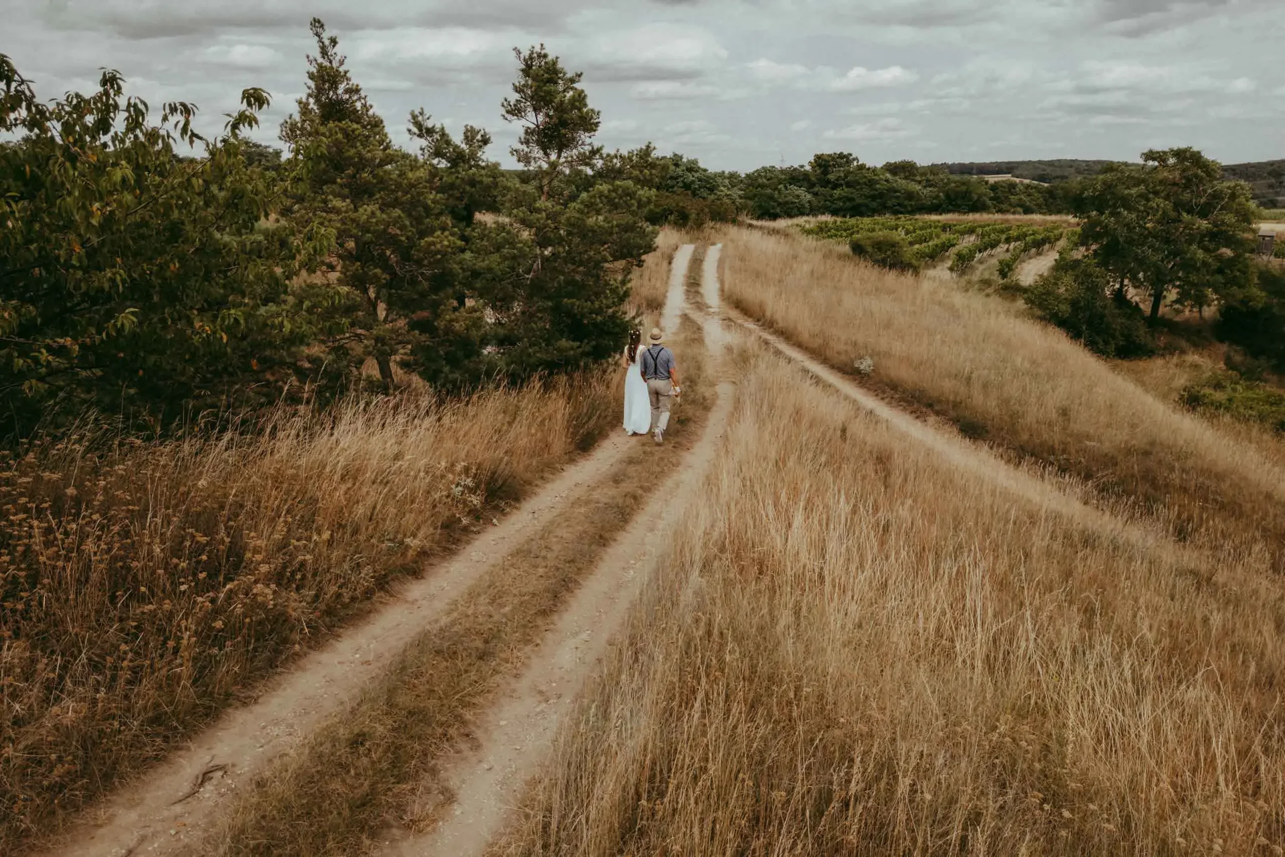 Zwei Menschen gehen auf einem unbefestigten Weg durch eine ländliche, hügelige Landschaft mit Gräsern, Bäumen und Weingärten, unter einem bewölkten Himmel.
