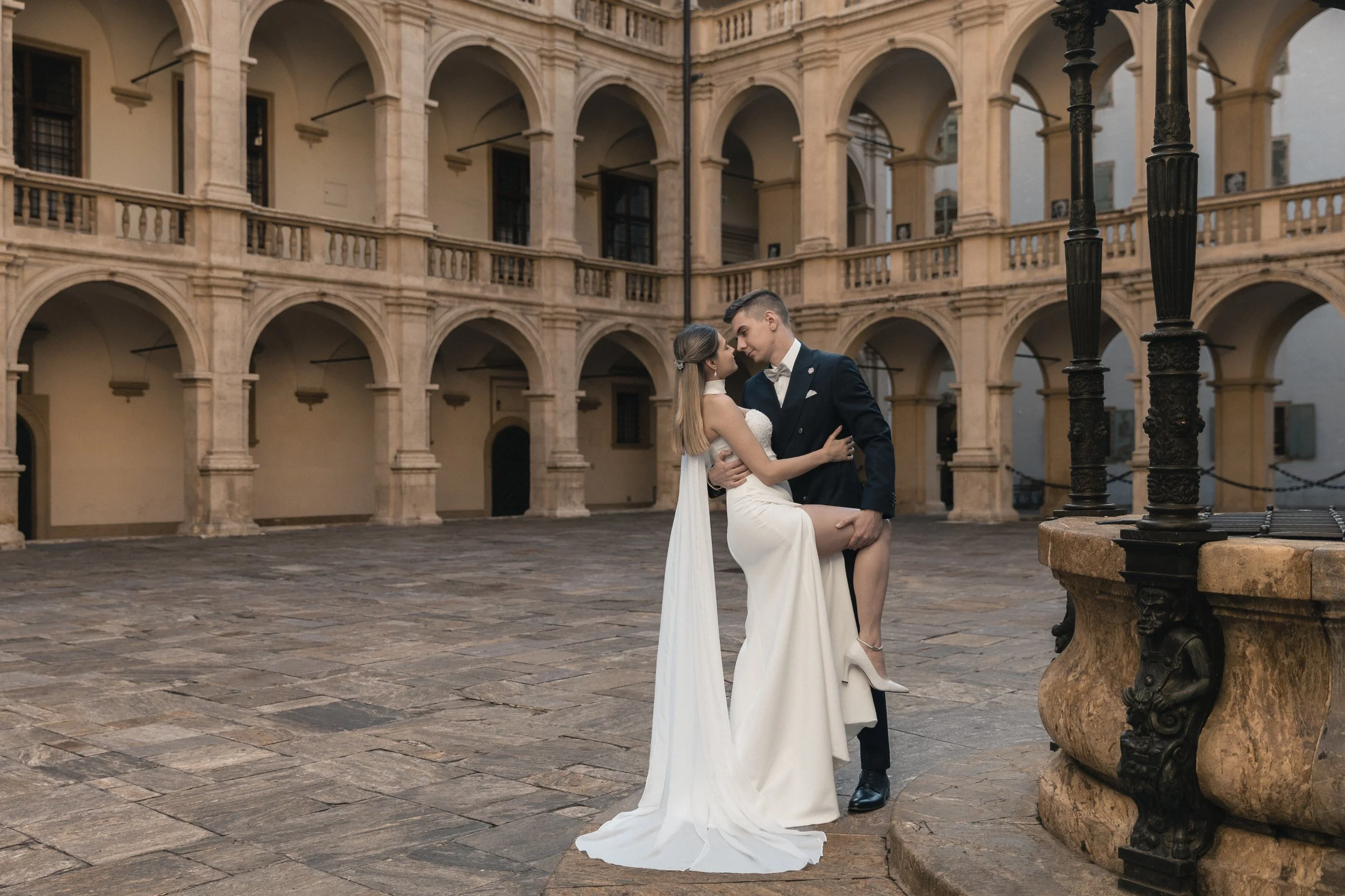 Ein Brautpaar umarmt sich in einem malerischen historischen Innenhof mit mehreren Arkaden und steinernen Säulen, bei Sonnenuntergang, romantische Stimmung, elegante Kleidung.