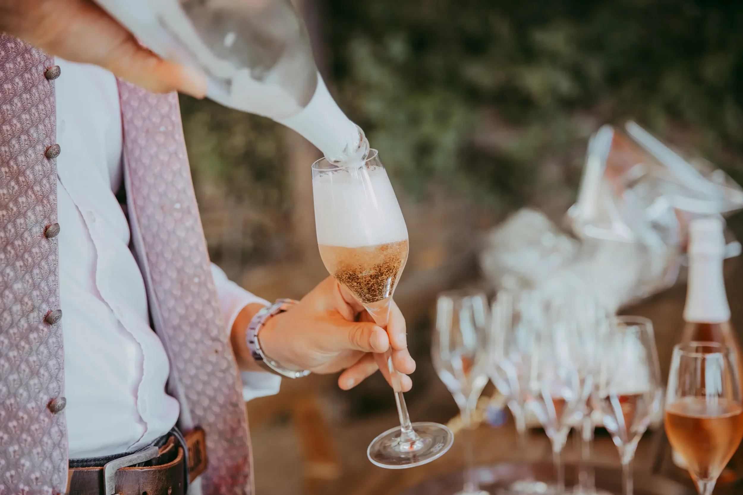 Eine Person in traditioneller Kleidung füllt ein Glas Champagner bei einer Feier im Freien.