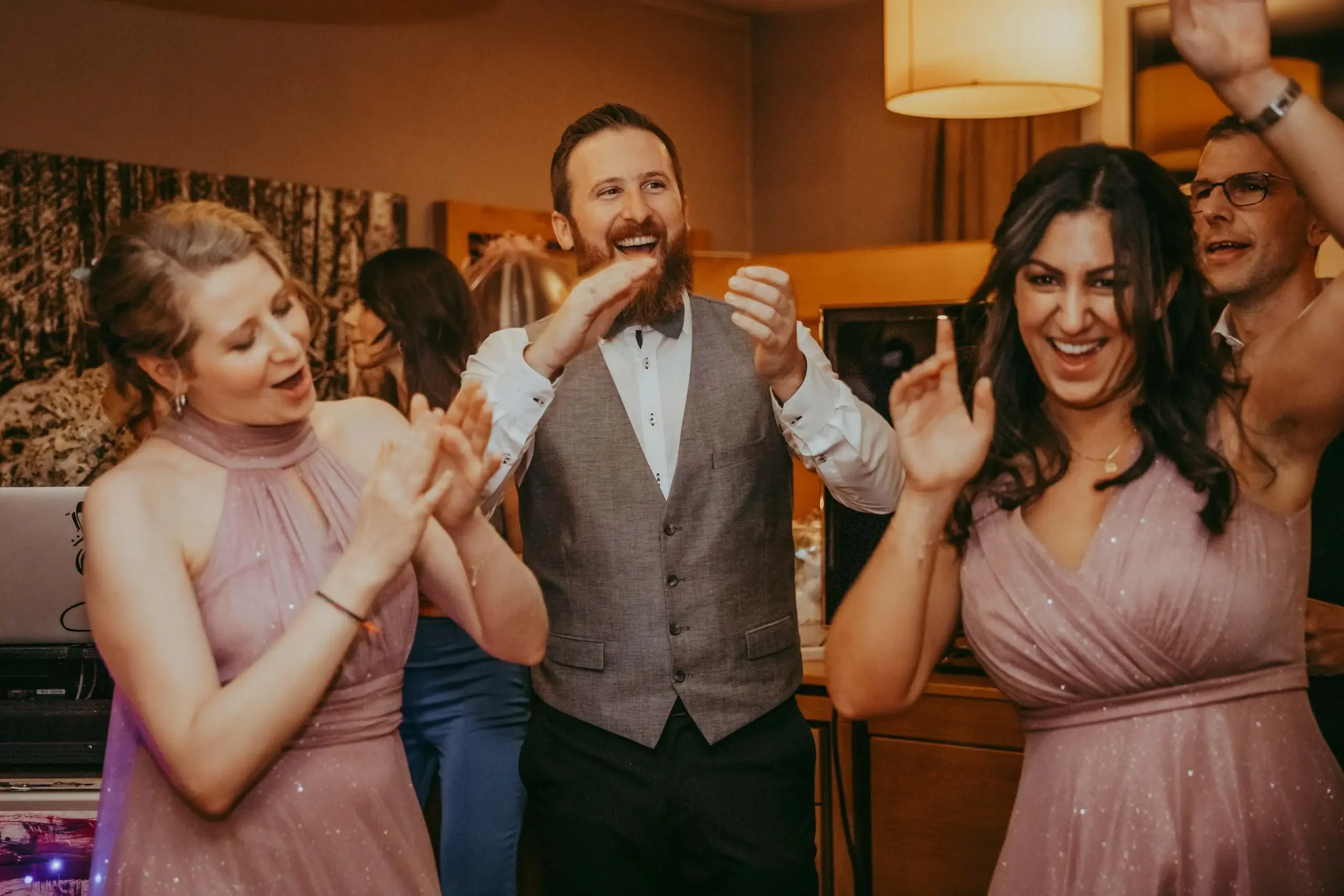 Gruppe von Menschen beim Feiern, lachen und Tanzen, zwei Frauen im pinken Kleid, ein Mann in Anzug mit Bart, im Hintergrund leuchtende Lampen.