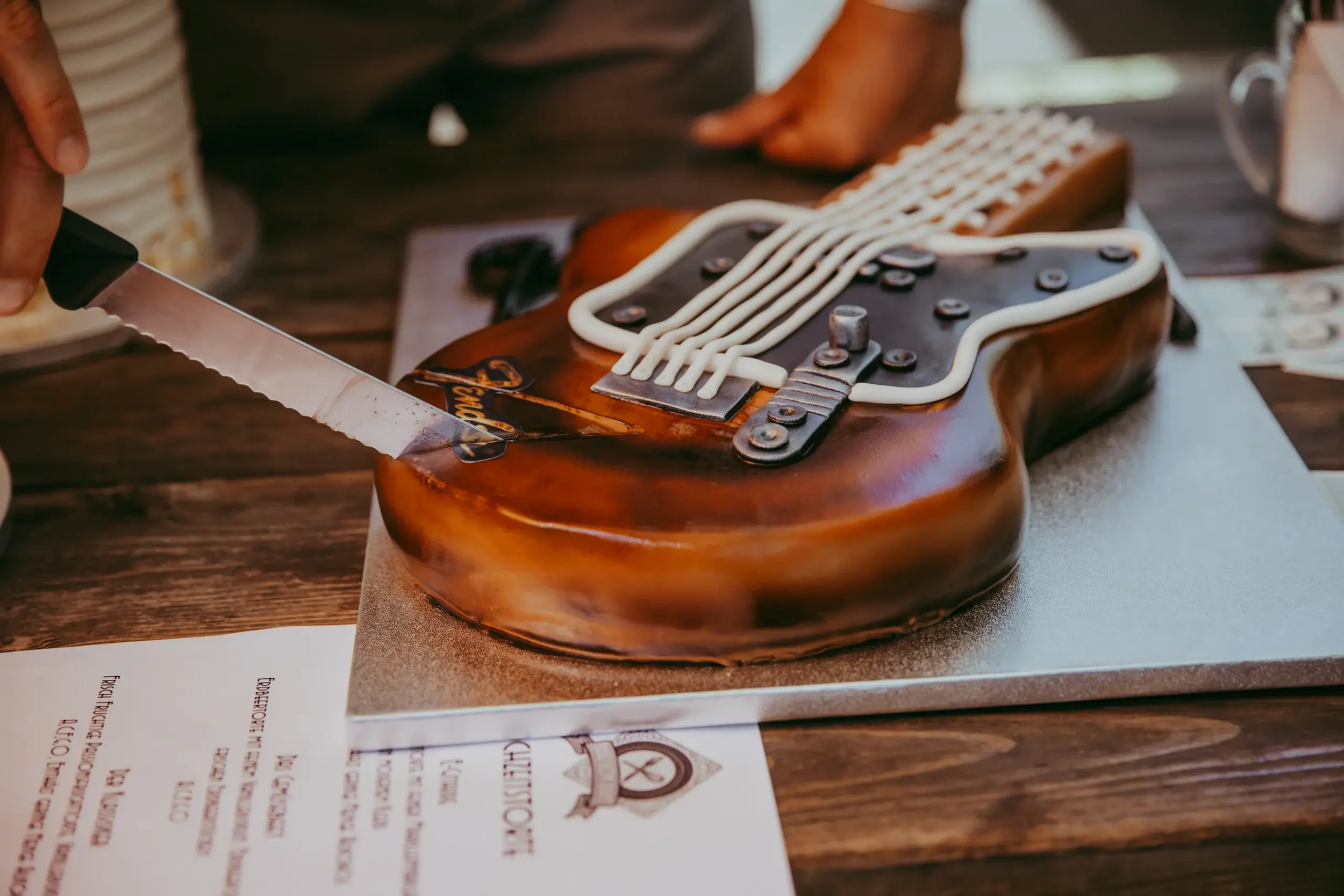 Ein Tortenmodell, das wie ein E-Gitarrenkörper gestaltet ist, mit weißen Zuckergusslinien und metallischen Schrauben zur Dekoration, auf einem Tisch.
