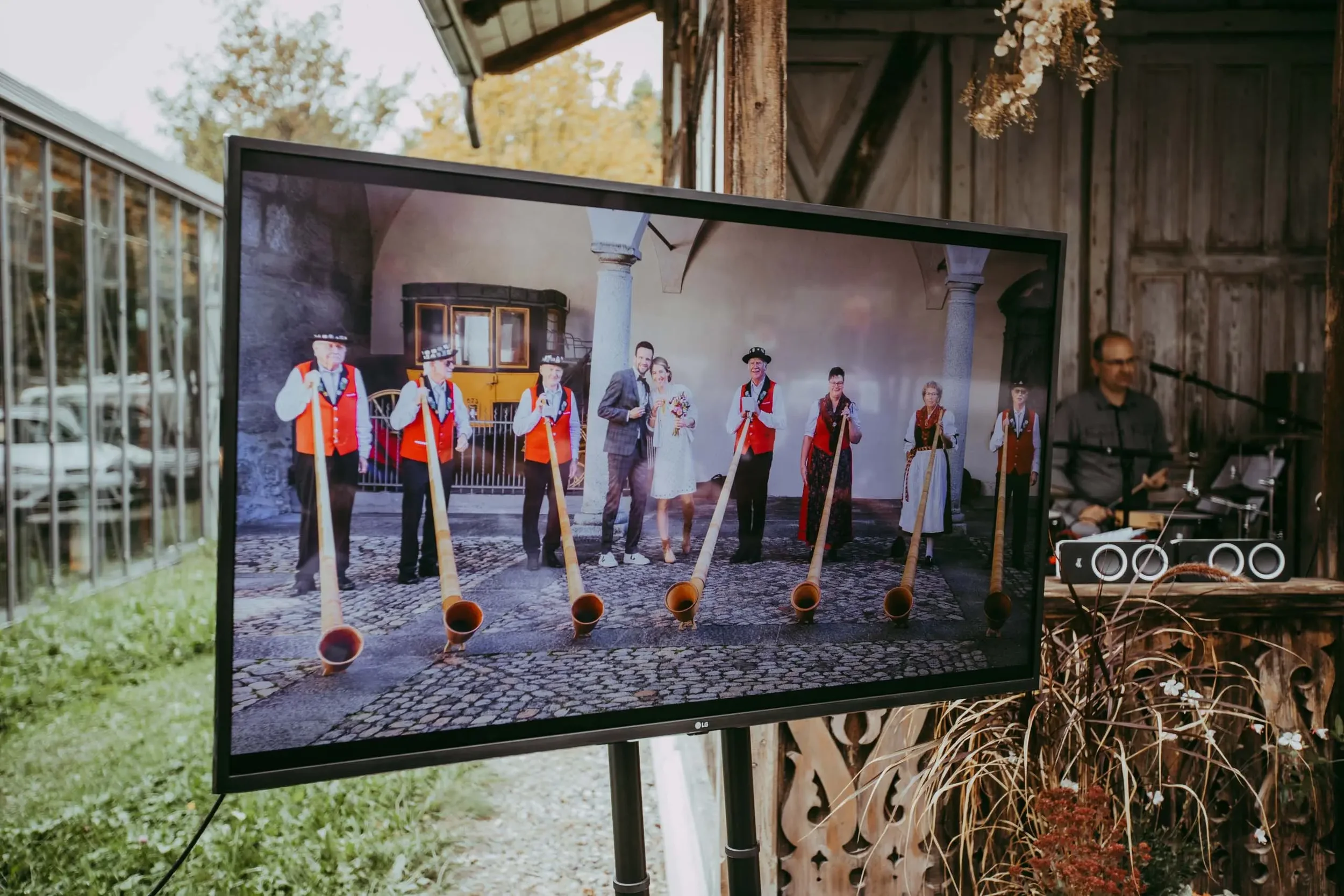 Foto eines Bildschirms mit einer Gruppe von Menschen in traditionellen Trachten, die auf einem Platz stehen, nahe einer alten Stadtmauer mit einem Gefährt im Hintergrund. Das Bild wird draußen gezeigt, umgeben von einem Holzzaun und Pflanzen.