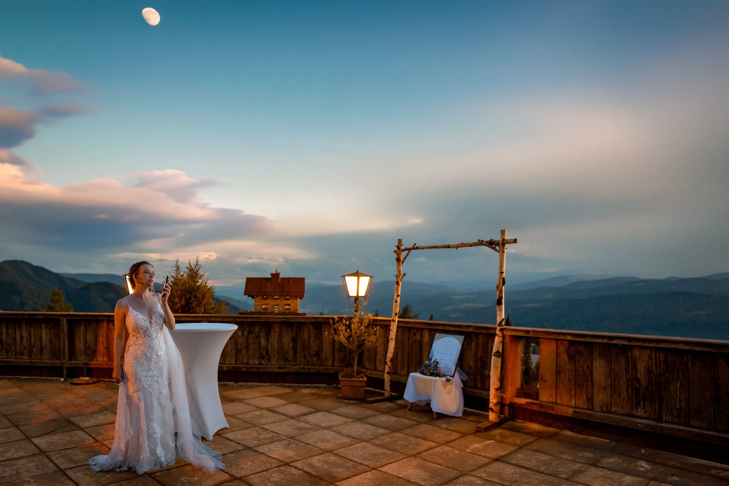 Eine Frau in einem weißen Hochzeitskleid steht auf einer Terrasse bei Sonnenuntergang, mit Bergen im Hintergrund, eine Laterne und ein Holzzaun sind sichtbar.