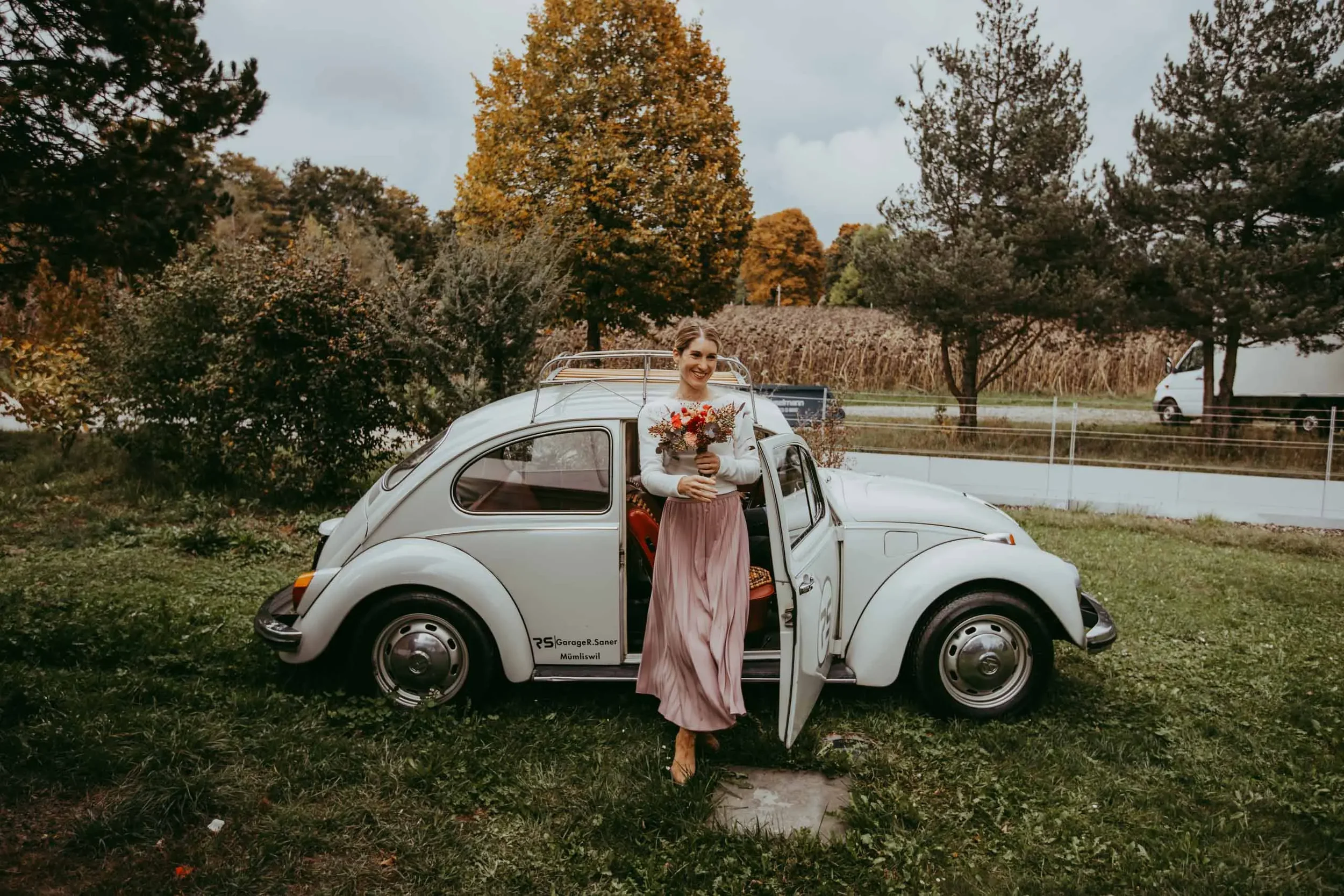 Eine lachende Frau steht vor einem weißen Autos in einer ländlichen Umgebung mit Bäumen im Herbst, hält einen Blumenstrauß in den Händen.