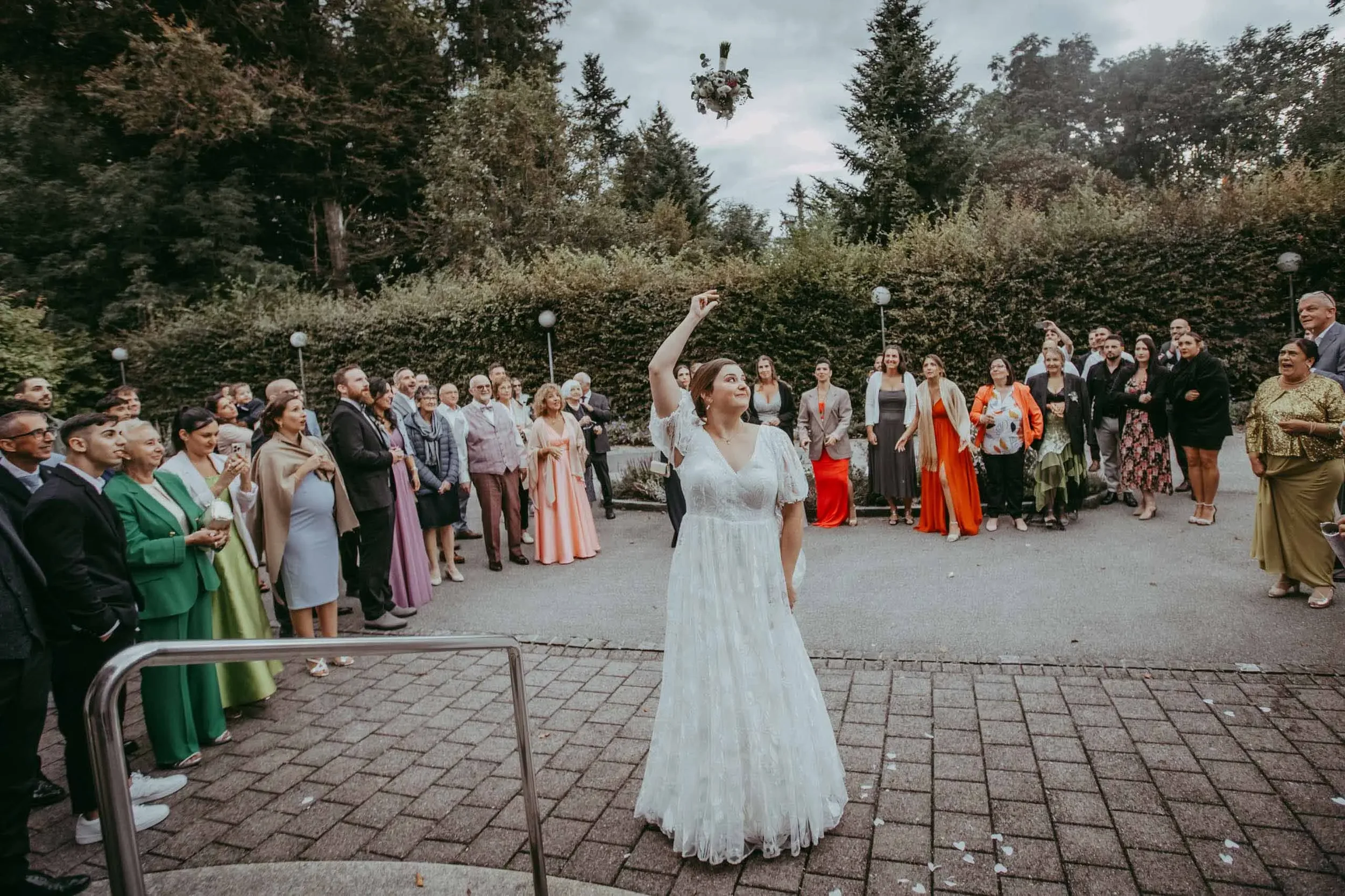 Eine Hochzeitsreportage mit einer Braut in einem weißen Kleid, die einen Brautstrauß in der Hand hält, während eine Gruppe von Menschen im Hintergrund bei einer Zeremonie im Freien zuschaut.
