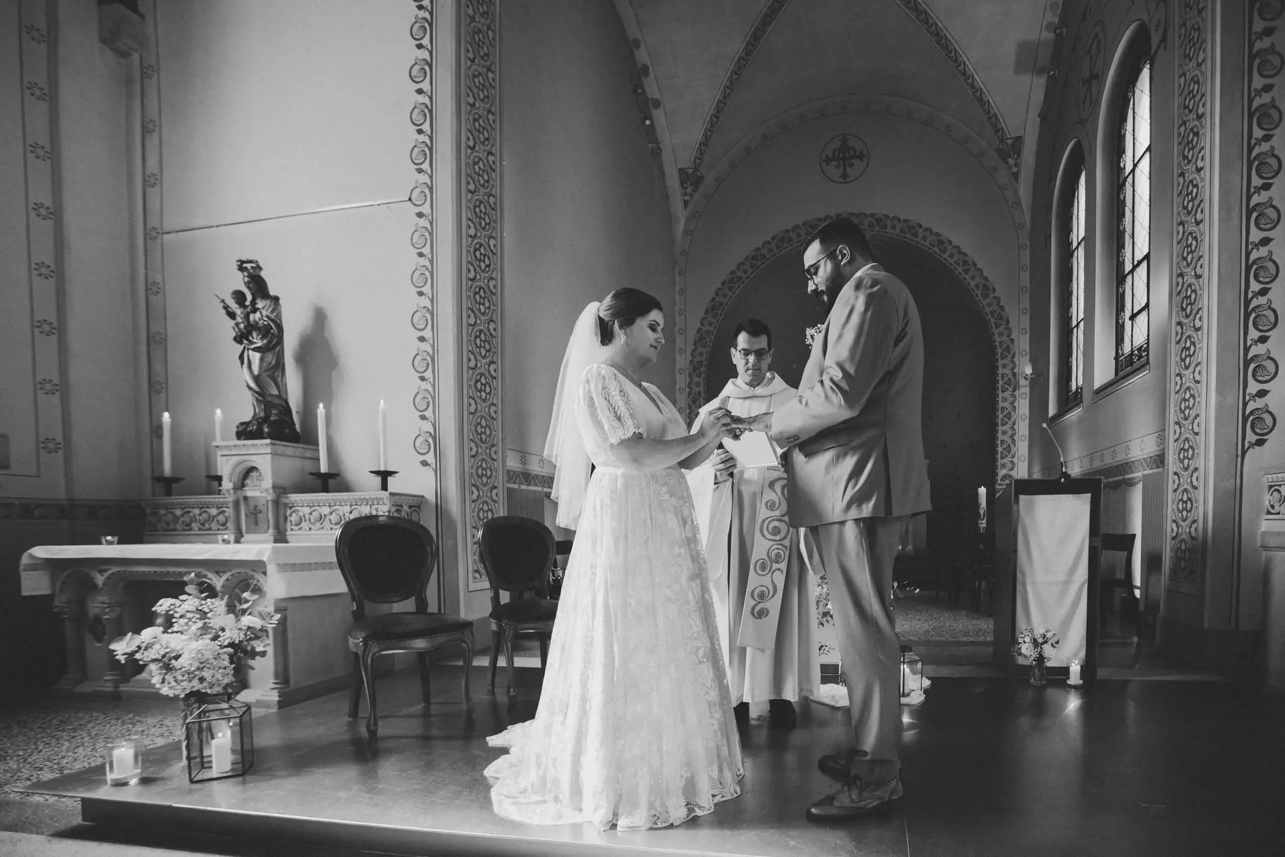 Eine Schwarz-Weiß-Fotografie einer Hochzeitszeremonie in einer Kirche. Ein brautpaar steht sich gegenüber, während der Priester dazwischen steht und die Trauung vollzieht. Das Paar besteht aus einer Frau in einem weißen Kleid und einem Mann in einem 