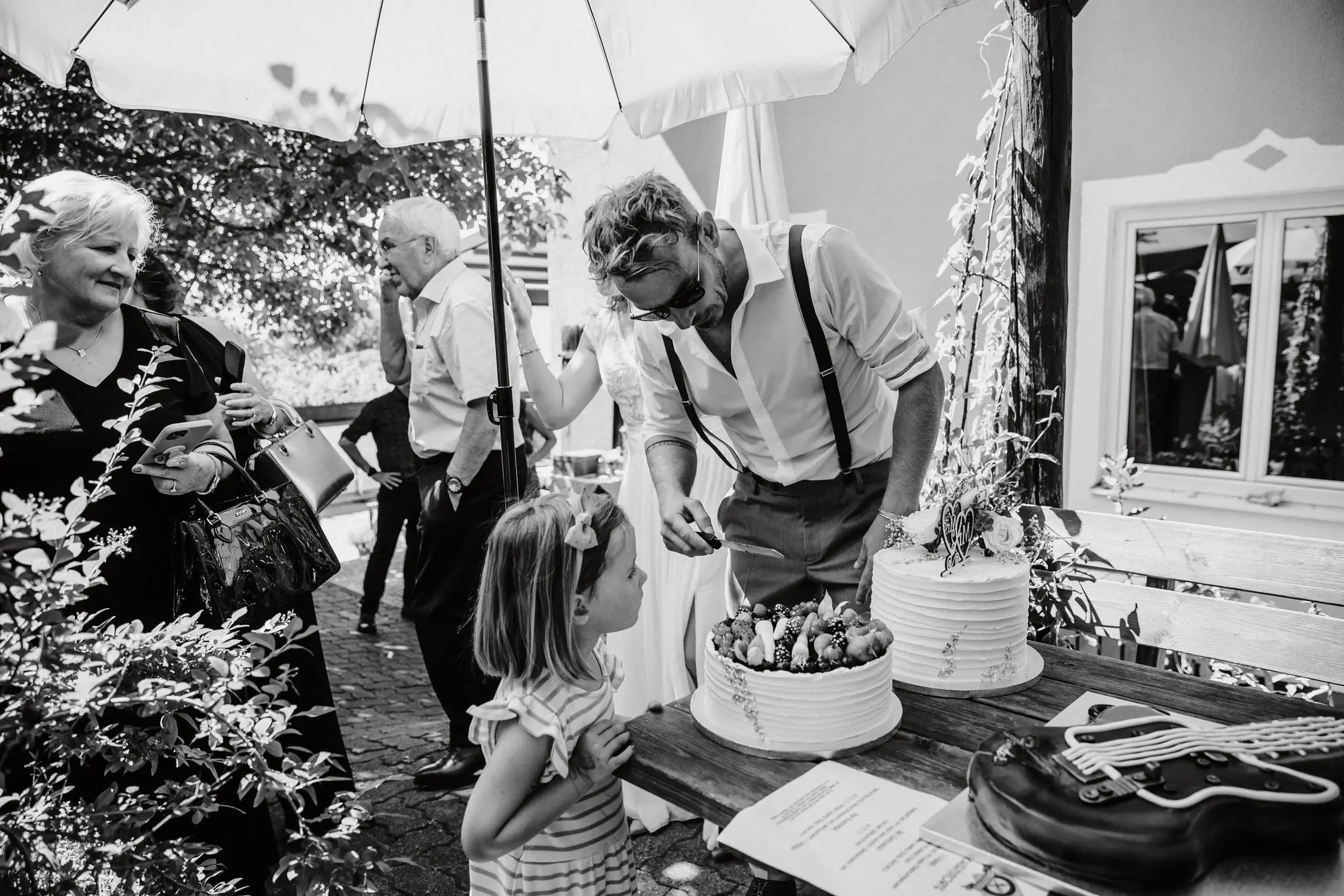 Menschen feiern draußen bei einer Hochzeit mit einem großen weißen Kuchen und Blumen, während ein Kind auf den Kuchen zeigt.