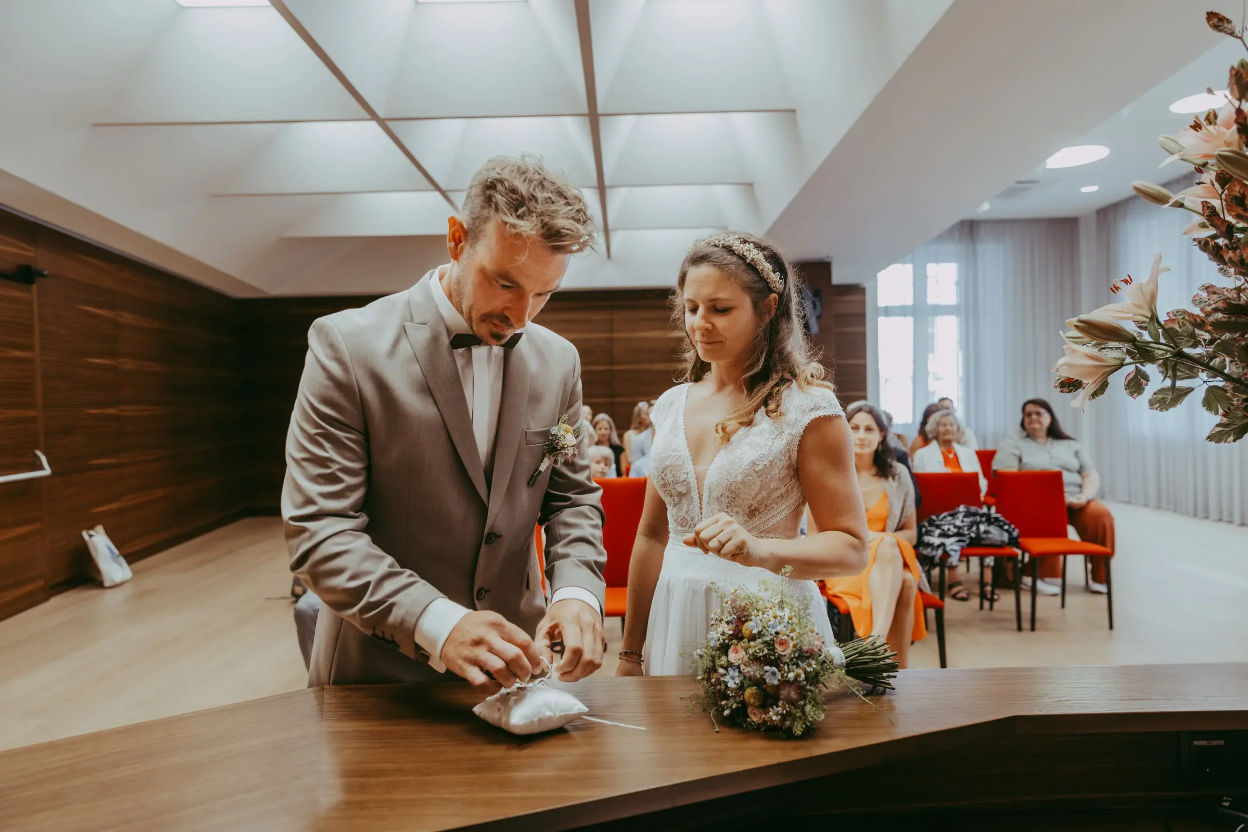 Ein Brautpaar bei einer Zeremonie, wahrscheinlich im Standesamt, während sie einen Ring an einem Tisch austauschen. Im Hintergrund sitzen Gäste und es gibt eine große Blumenan arrangement auf der rechten Seite.