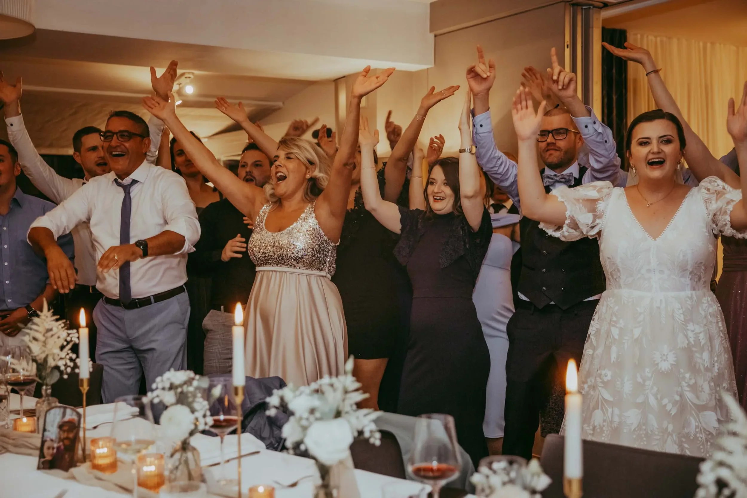 Menschen feiern fröhlich bei einer Hochzeit, die Hände in die Luft gestreckt, im festlich dekorierten Raum.