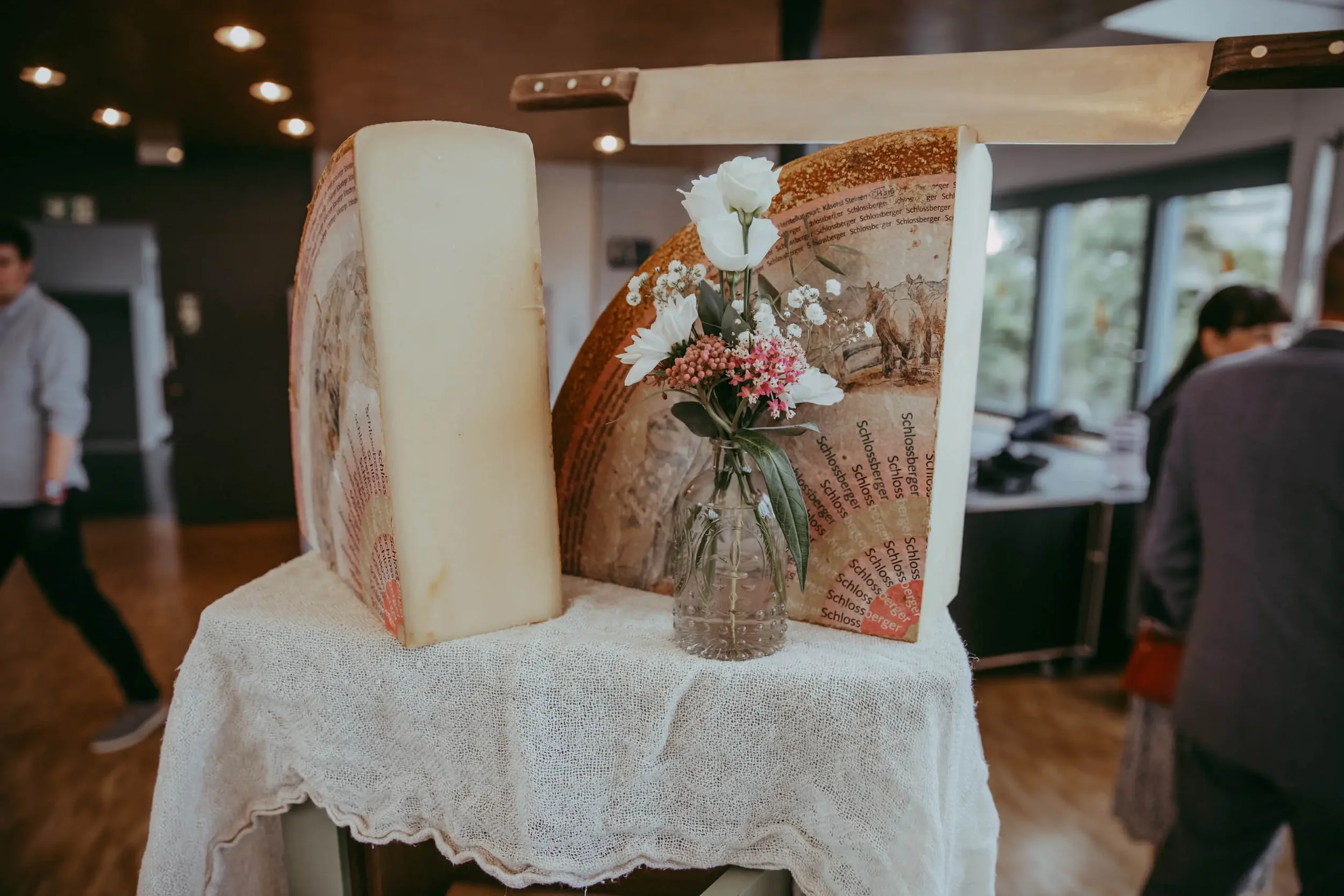 Ein Tablett mit verschiedenen Käsesorten, darunter ein großer Käseblock, dekoriert mit einem kleinen Blumenstrauß in einer Glaskaraffe, im Hintergrund eine helle Raumumgebung mit Fenstern und mehreren Menschen.