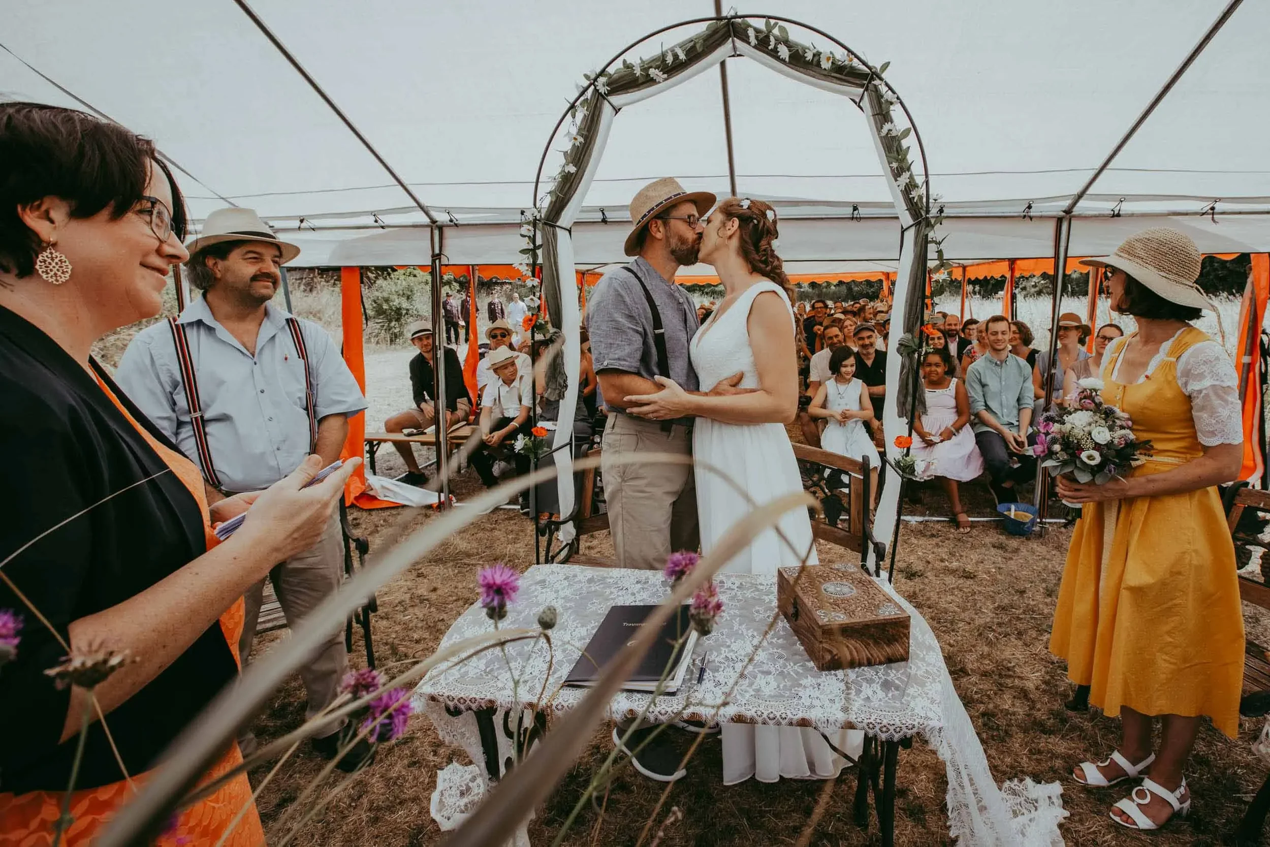 Ein Paar küsst sich während ihrer Hochzeit unter einem festlichen Zelt im Freien, umgeben von Gästen.