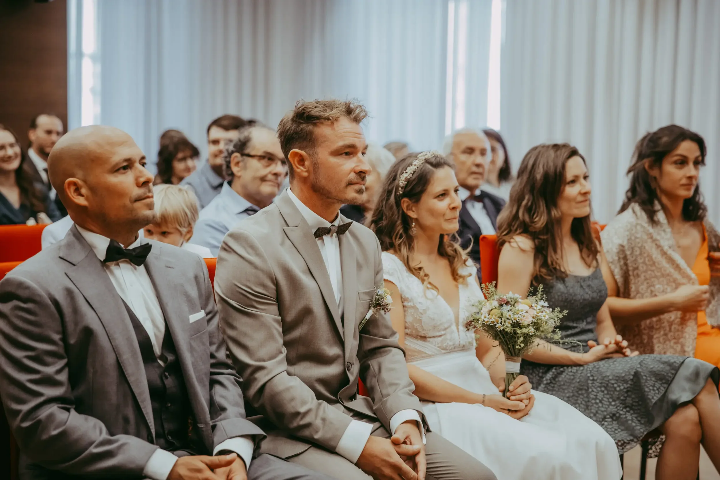 Hochzeitspaar sitzt unter den Gästen bei einer standesamtlichen Trauung, umgeben von Verwandten und Freunden in einem hellen Saal.