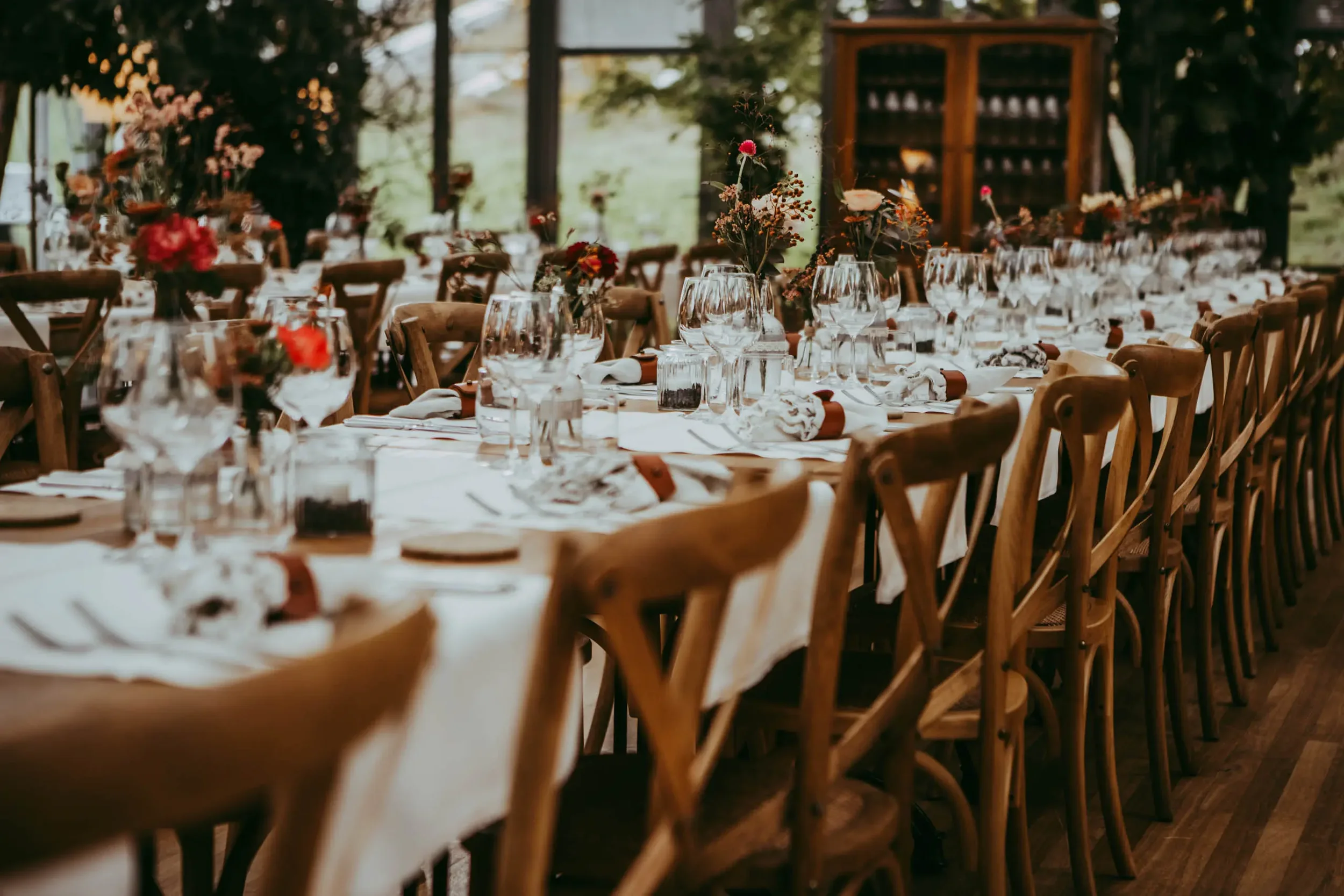 Ein festlich gedeckter Tisch in einem rustikalen Raum mit Holzstühlen, Blumenarrangements und Gläsern, vermutlich für eine Hochzeit oder Feier vorbereitet.