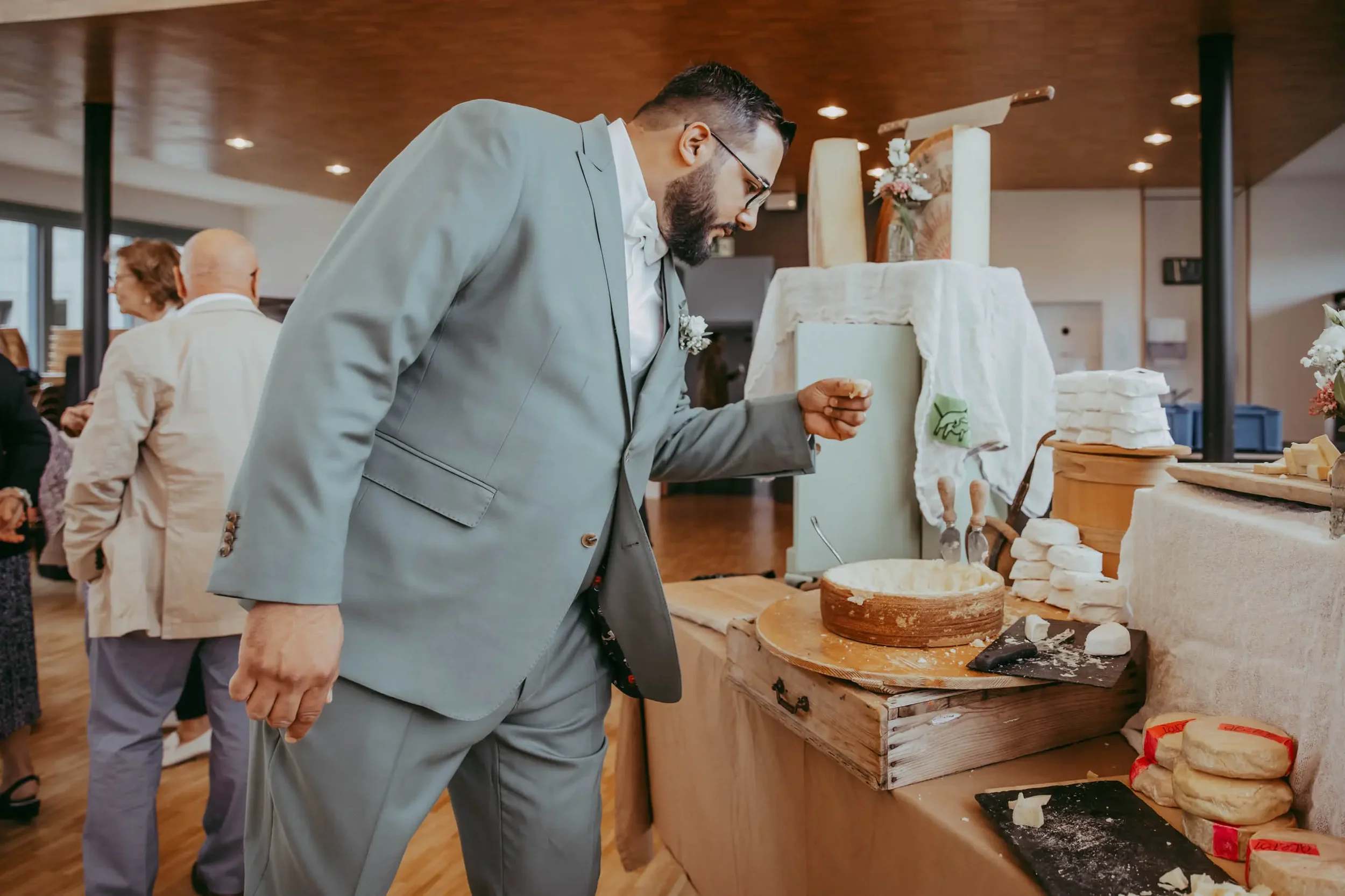 Ein Mann in einem grauen Anzug mit Brille betrachtet Käse- und Käseähnliche Produkte auf einem Tisch in einem Raum mit Holzdecke. Es gibt mehrere Personen im Hintergrund.