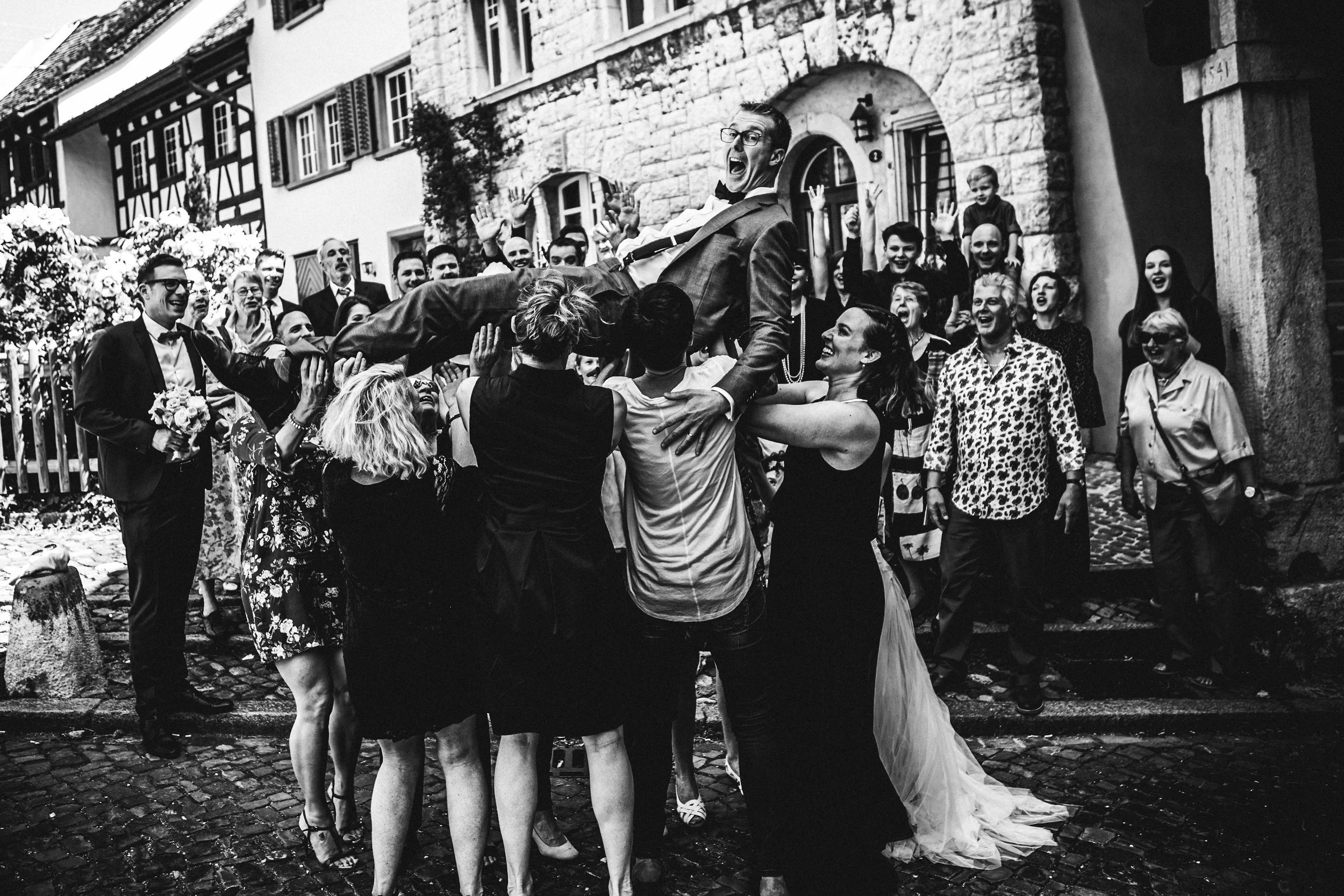 Eine Gruppe von Menschen beim Hochzeitsfest, bei dem ein Mann in formeller Kleidung hochgehoben wird, während er lachend und mit offenen Armen in einer eleganten Straße mit alten Gebäuden steht.