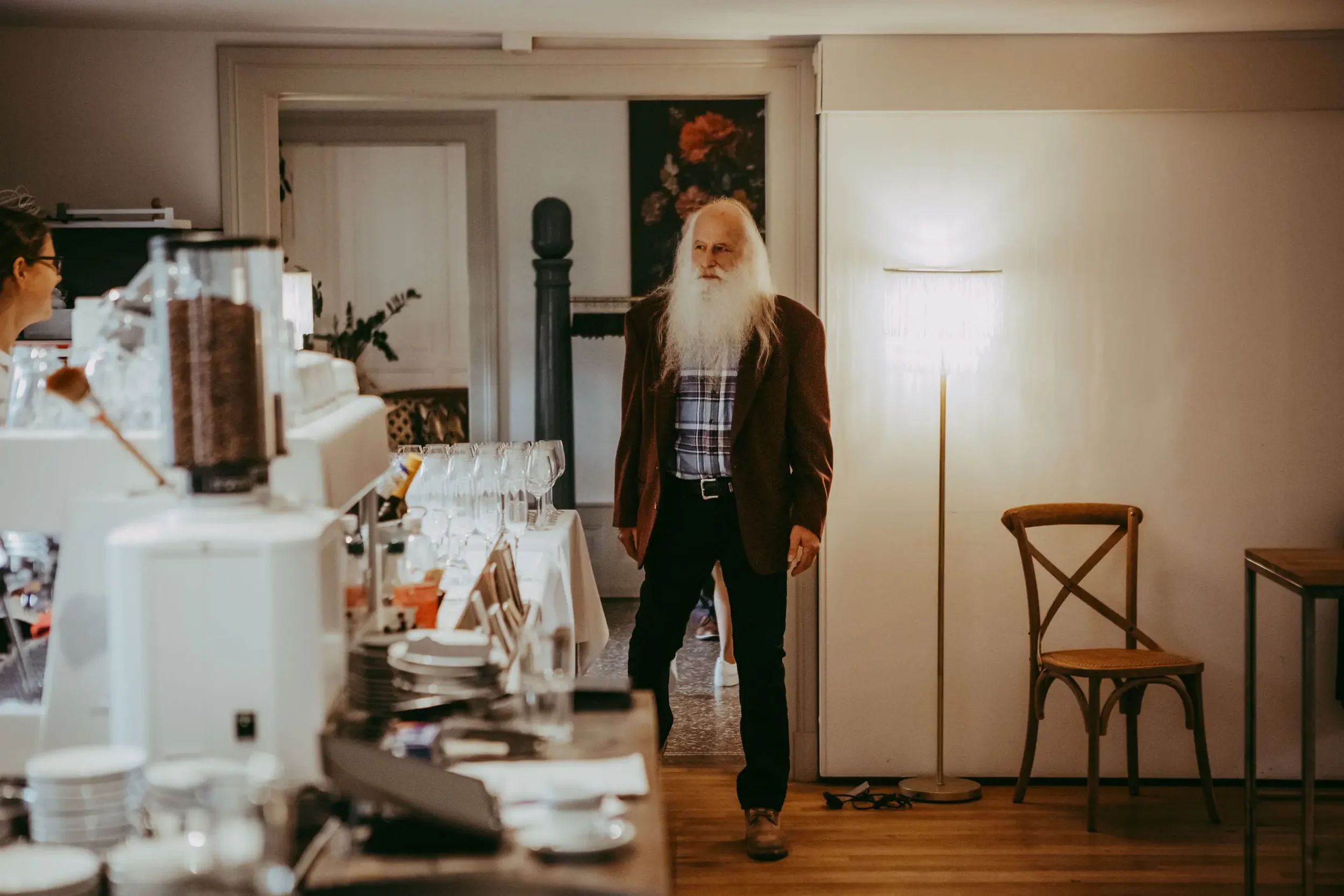 Ein älterer Mann mit langem weißem Bart und langen weißen Haaren, trägt eine braune Jacke und ein kariertes Hemd, steht in einem Raum vor einer Tür, im Hintergrund eine Stehlampe und eine Wand mit einem Blumenbild.