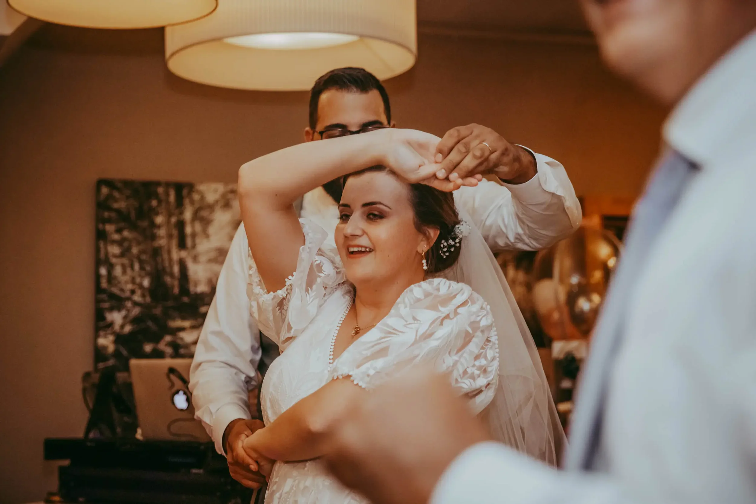 Ein Mann macht eine Hochzeitszeremonie mit einer Braut, die eine weiße Hochzeitskleidung trägt, während sie lachen und sich umarmen, im Hintergrund eine Wohnzimmer-Umgebung mit warmem Licht.