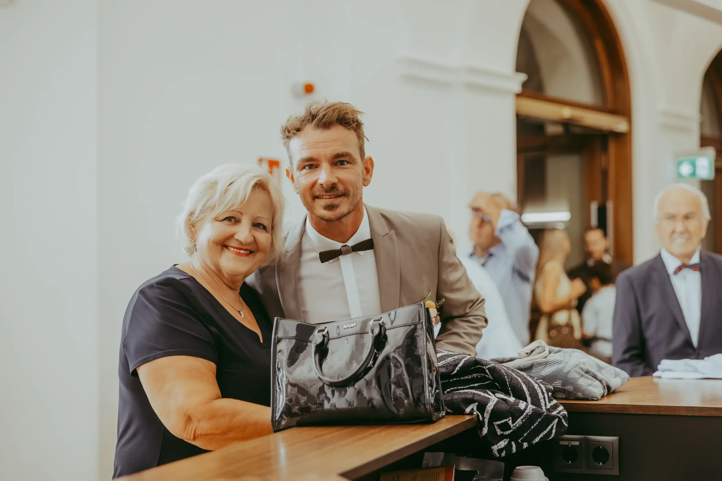 Ein älterer Frau und ein junger Mann posieren zusammen bei einer Veranstaltung. Im Hintergrund sind andere Personen zu sehen, die sich unterhalten.