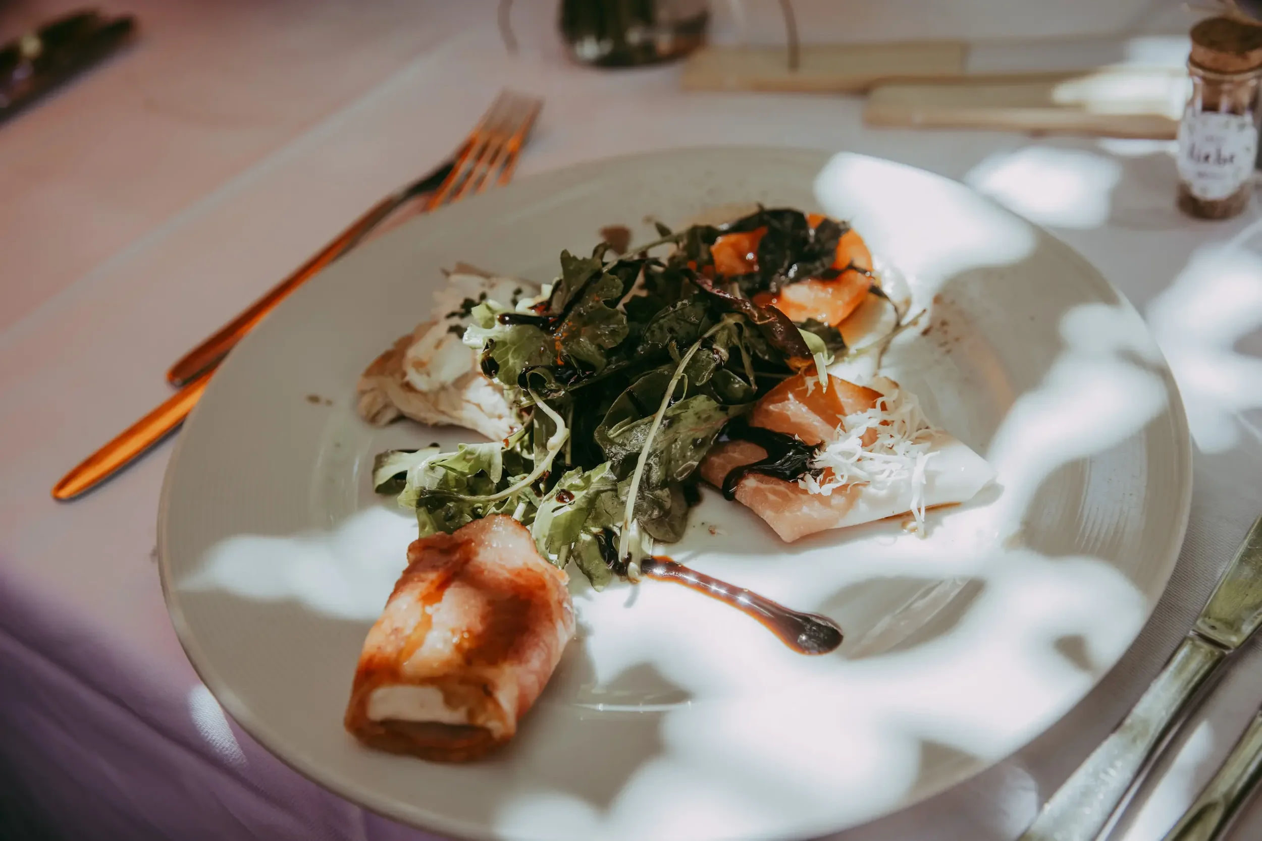 Auf einem weißen Teller befinden sich ein Salat mit dunklen Blättern, ein Stück geräucherten Fisch, einige Blätter und eine dunkle Soße. Daneben liegt eine knusprige, goldbraune Bratkartoffel. Im Hintergrund sind Besteck und eine kleine Flasche zu se