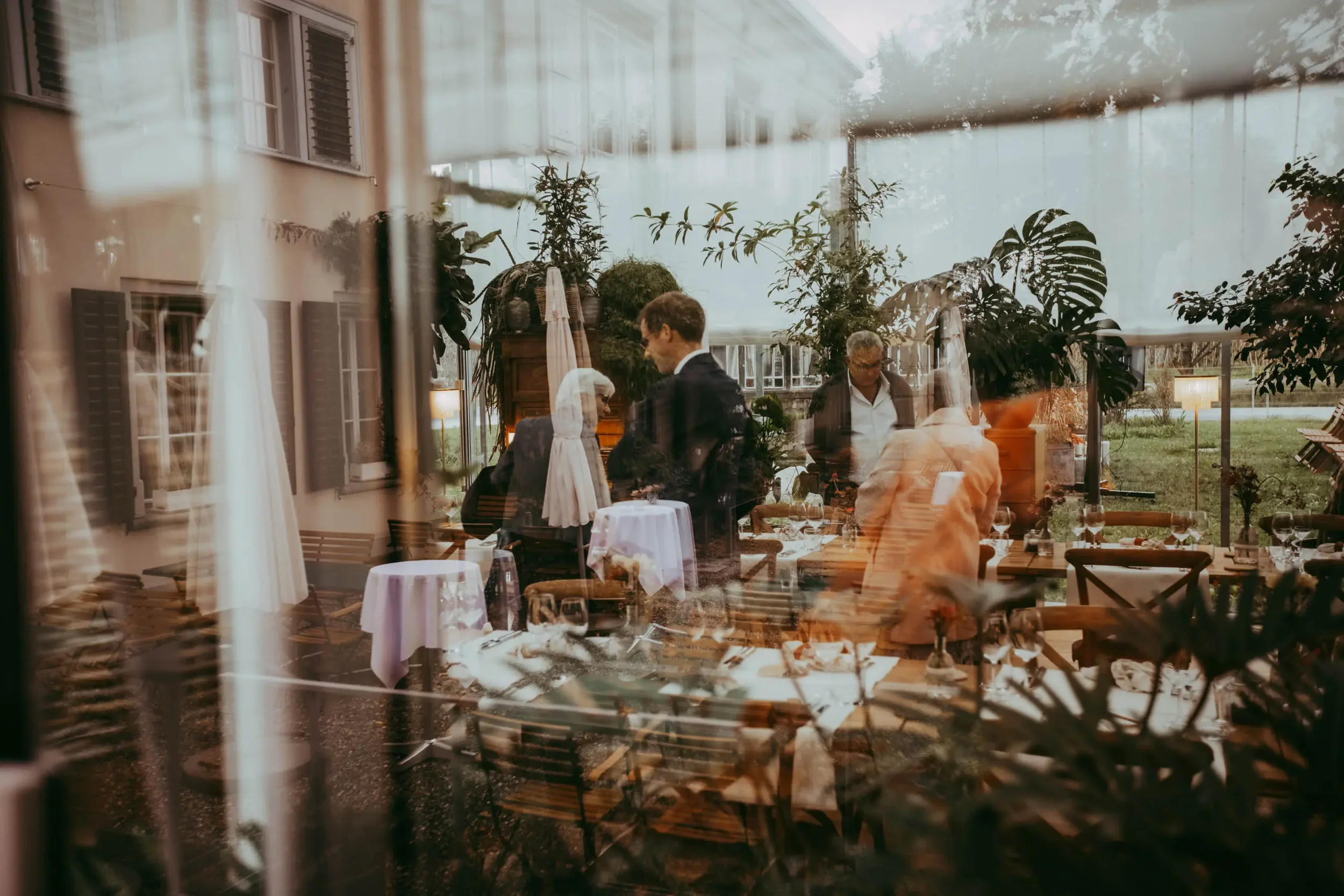 Blick durch eine Fensterfront auf eine Gartenparty im Innenhof mit mehreren Tischen und Gästen.