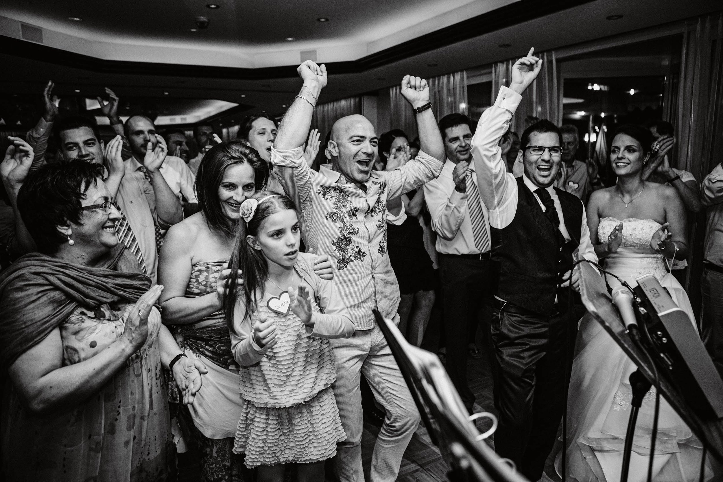 Menschen feiern bei einer Tanzparty, einige mit erhobenen Armen, in einem festlich dekorierten Raum, schwarz-weiße Aufnahme.