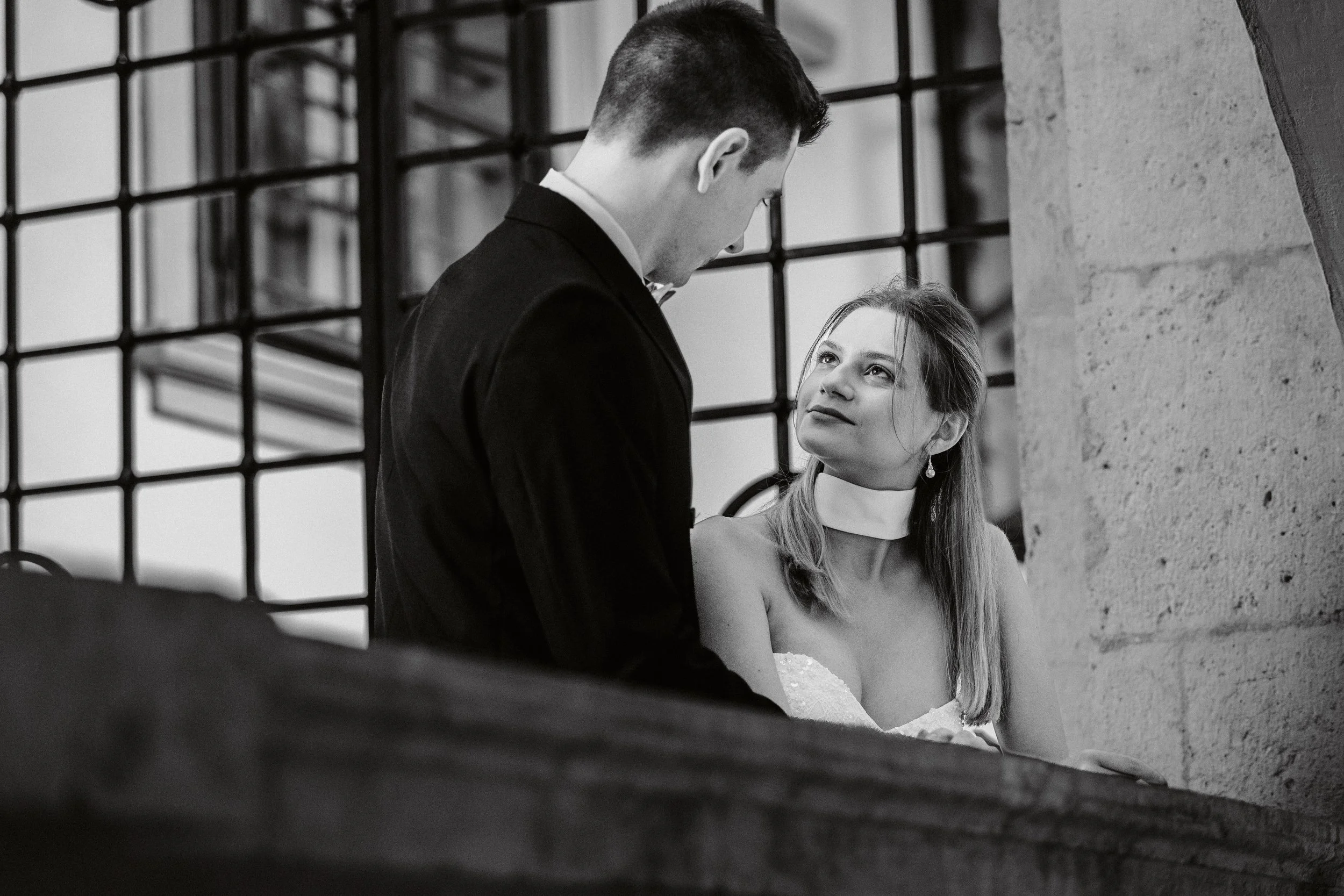 Ein Mann und eine Frau stehen sich gegenüber an einem Rand eines Raumes, der durch ein Gitter im Hintergrund sichtbar ist. Das Bild ist schwarz-weiß und zeigt einen intimen Moment zwischen den beiden.