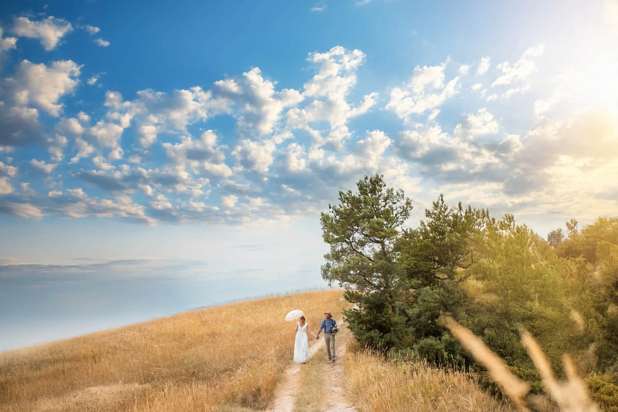 Ein Paar geht Hand in Hand auf einem Pfad durch eine goldene Wiesenlandschaft, während die Frau einen weißen Sonnenschirm trägt und der Mann einen Blumenstrauß hält. Im Hintergrund gibt es einen großen Baum und den klaren Himmel mit vereinzelten Wolk