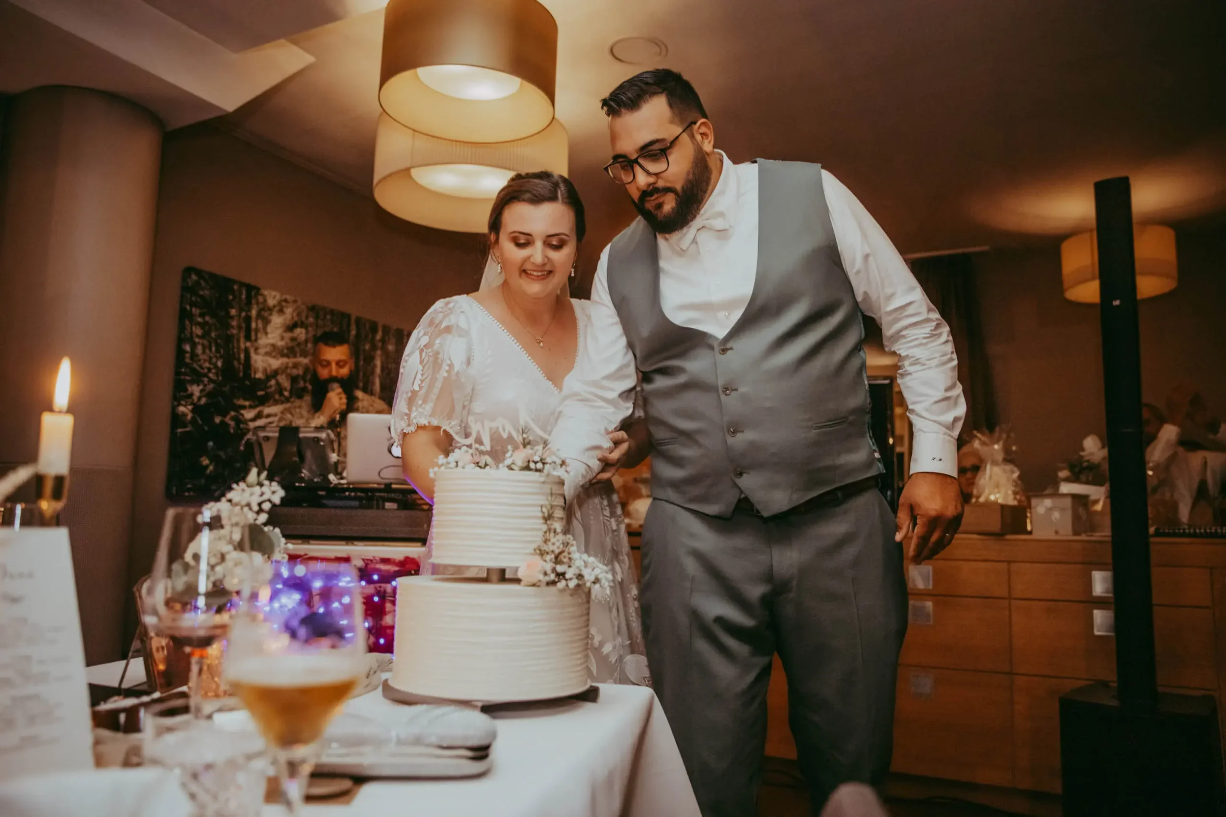 Ein Brautpaar schneidet zusammen eine Hochzeitstorte bei einer Feier mit Kerzen und Dekorationen im Hintergrund.