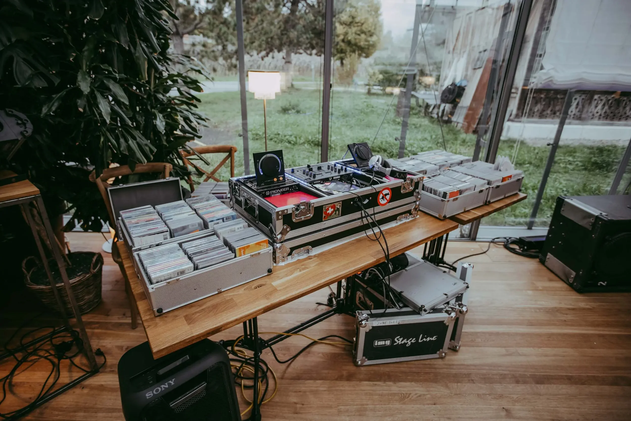 Ein DJ-Setup mit Plattenspielern, einem DJ-Mixer, CDs, Lautsprechern und einer Bühne, im Inneren eines Glasanbaus mit Blick auf einen Garten mit Bäumen und Rasen.