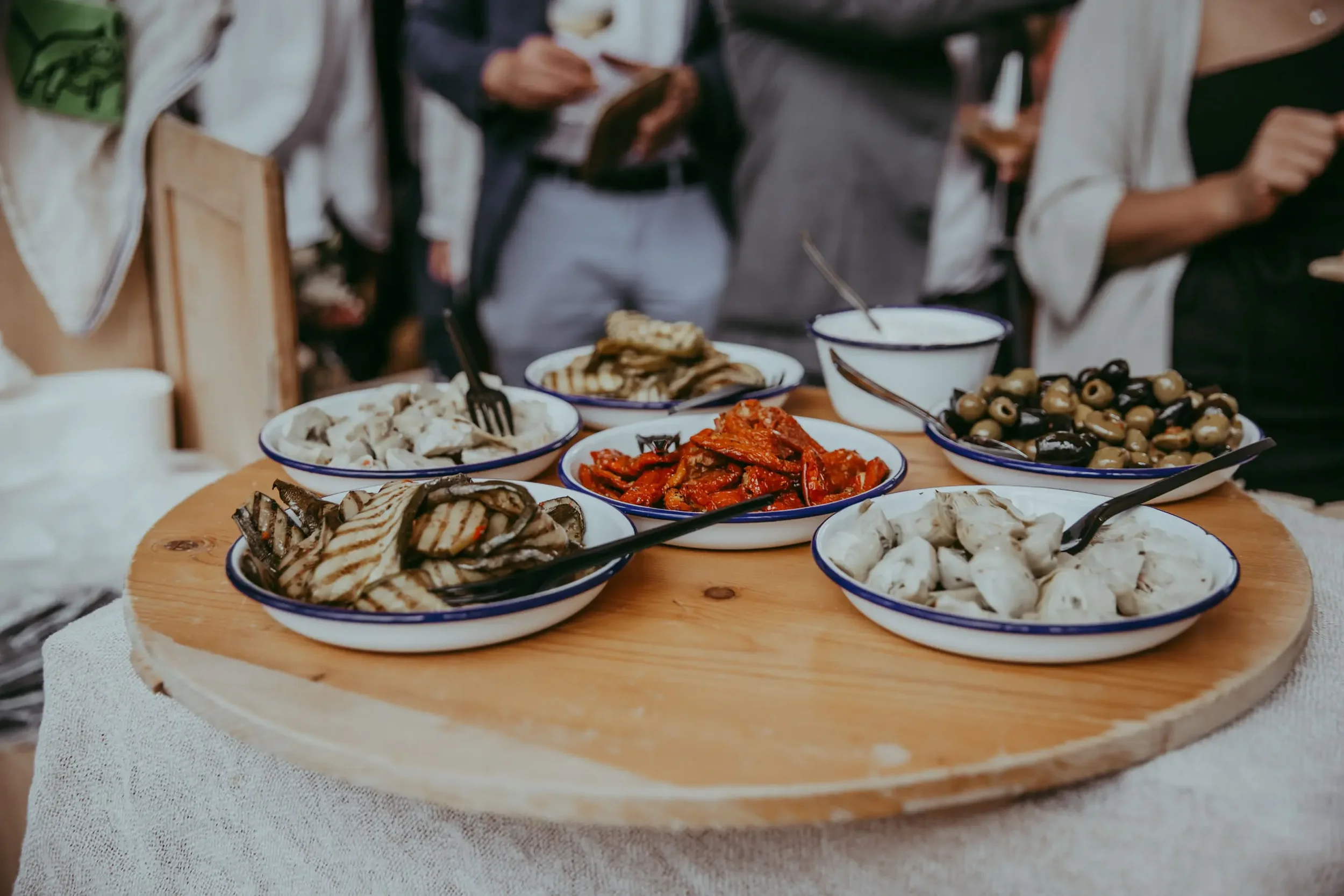 Verschiedene mediterrane Vorspeisen in blauen und weißen Keramiktellern auf einem Holzteller. Es gibt gegrilltes Gemüse, Oliven, Käse und Antipasti, auf einem festlichen Esstisch.