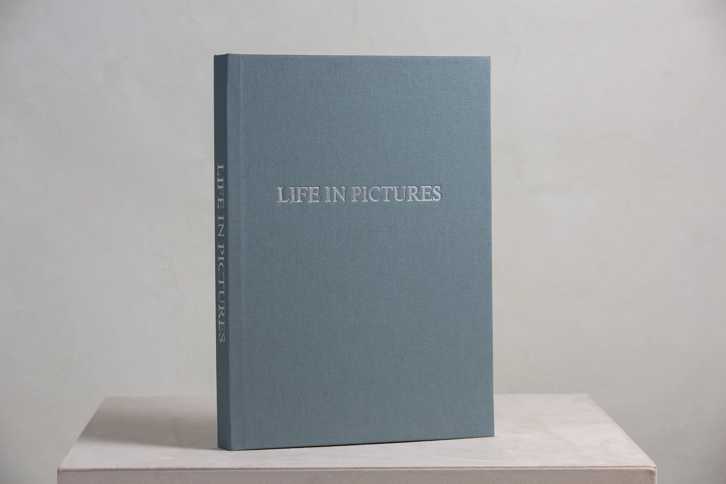 Ein blaues Buch mit dem Titel 'Life in Pictures' steht auf einer weißen Ablage vor einem hellgrauen Hintergrund.