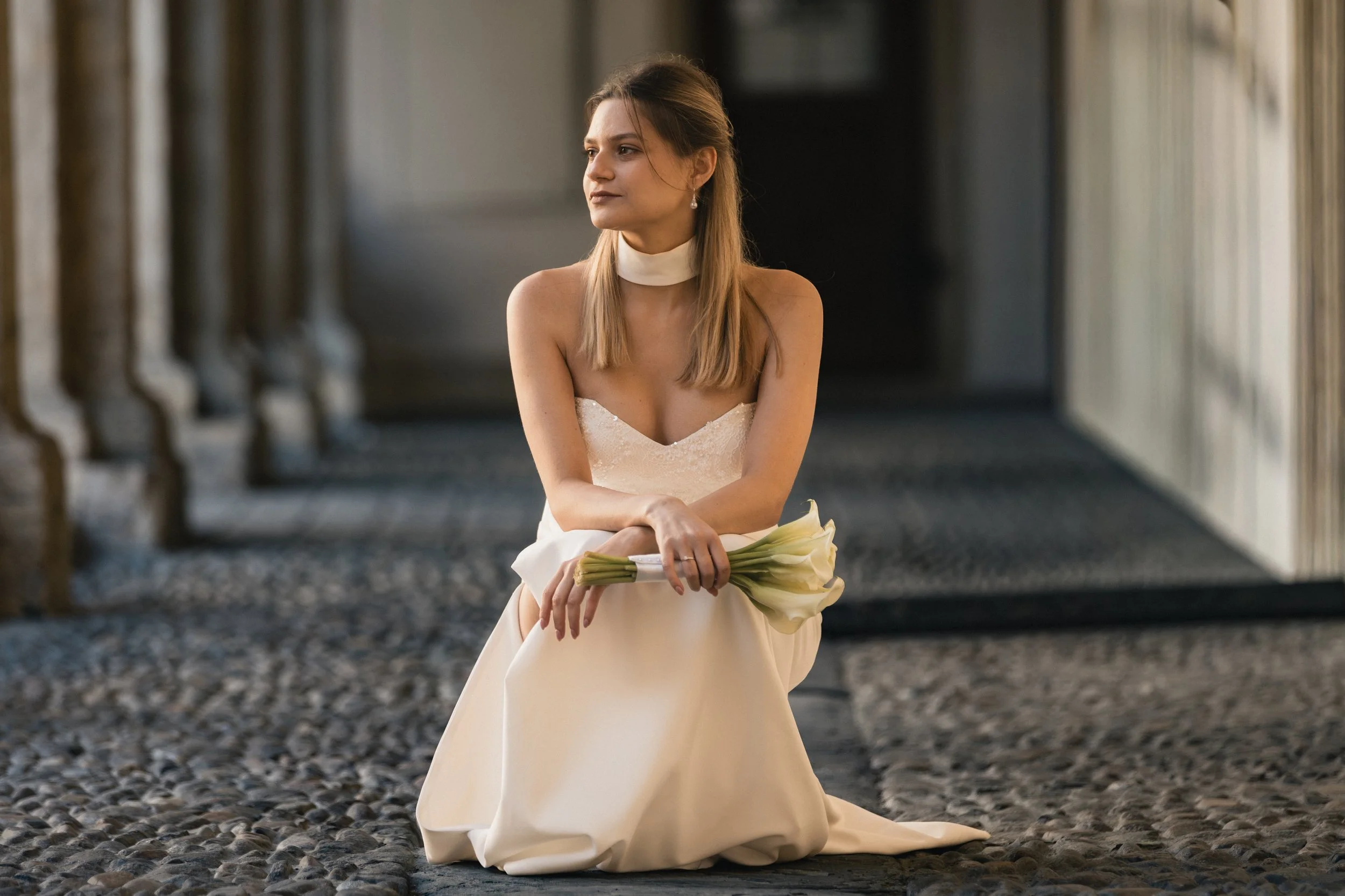 Eine junge Frau in einem weißen Brautkleid sitzt im Freien, hält einen Blumenstrauß in den Händen und schaut nach links.