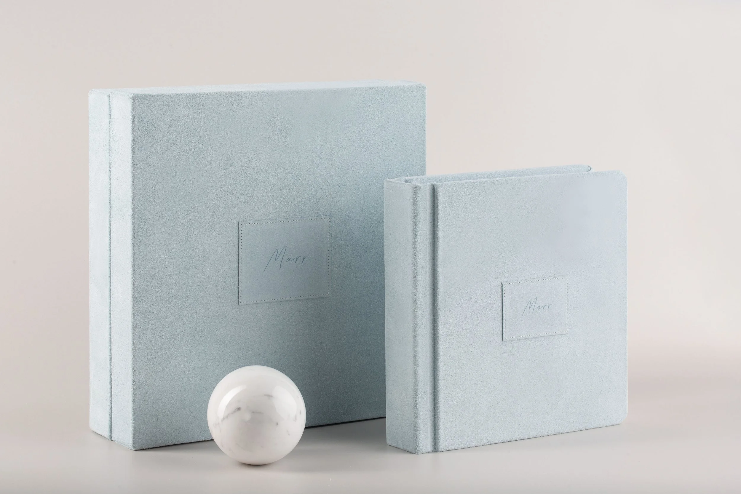 Zwei Geschenkboxen in Pastellblau mit Woll-Textur, eine große und eine kleinere, neben einer weißen Marmor-Kugel.